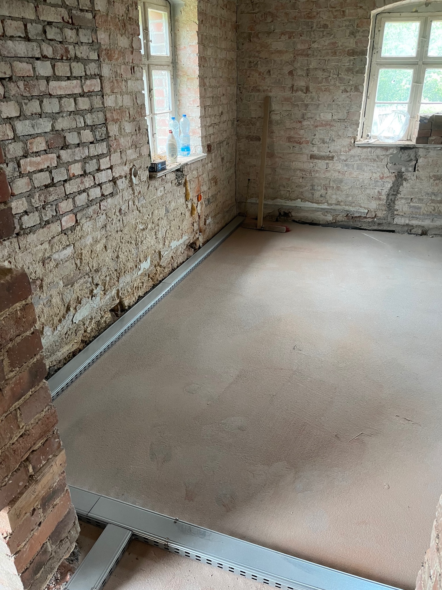 Surowy remont pokoju: odsłonięta cegła, świeżo wylana podłoga z listwami przypodłogowymi, okna z widokiem na zieleń. Widoczne ślady wcześniejszych prac budowlanych.