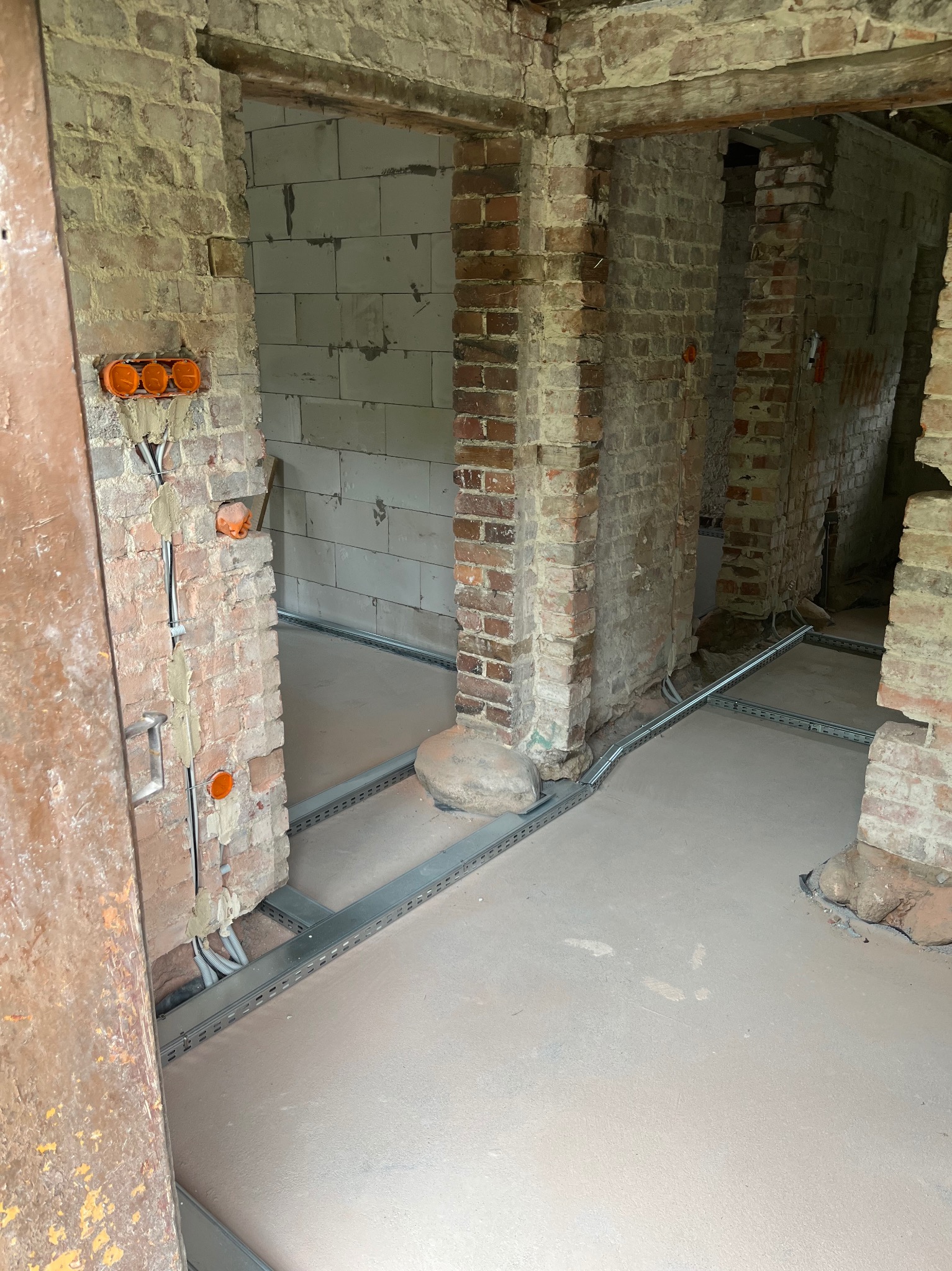 Surowy stan wnętrza z instalacją elektryczną: kable w korytach podłogowych, puszki w murze. W tle widoczne surowe ściany z cegły i betonu komórkowego.