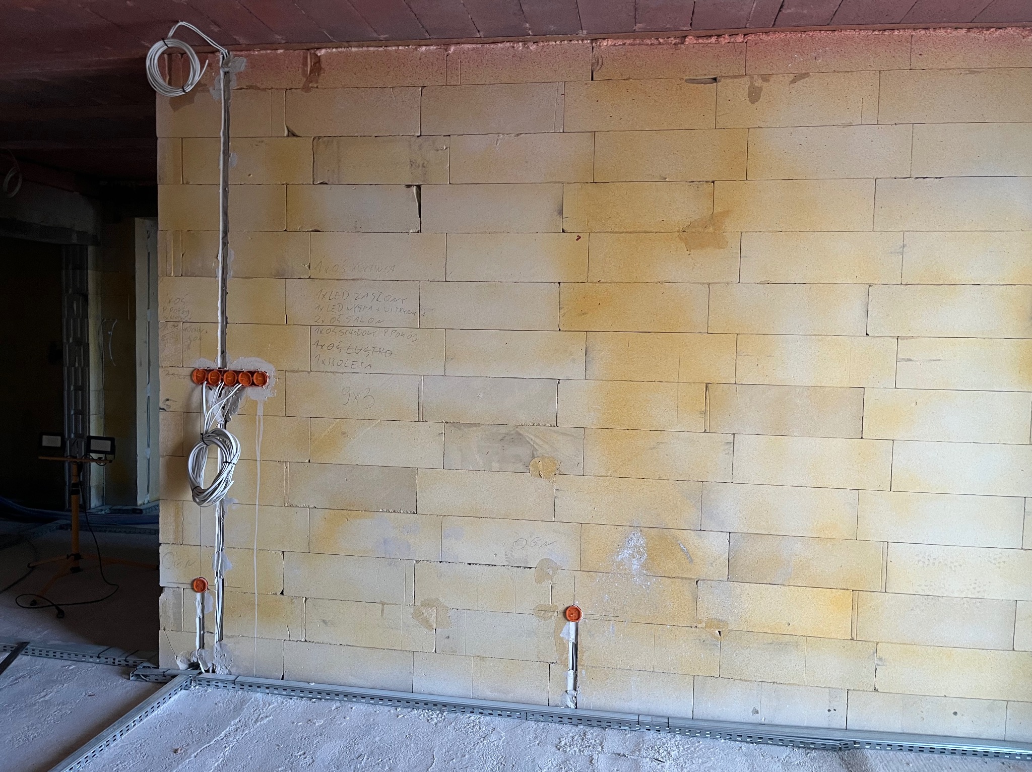 Ściana z instalacją elektryczną w trakcie montażu: kable, puszki, listwy przypodłogowe. Na ścianie notatki dotyczące rozmieszczenia punktów świetlnych.