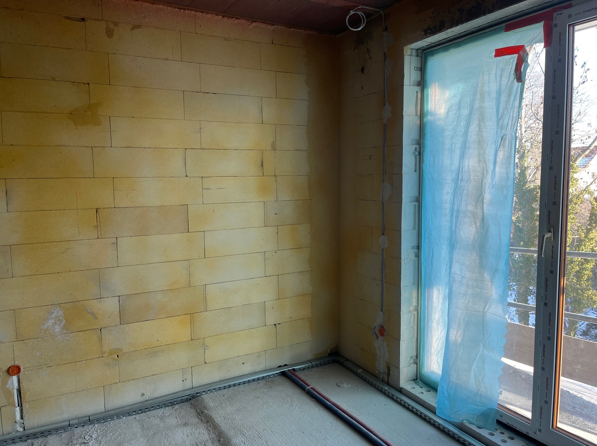 Wnętrze w trakcie prac instalacyjnych: ściana z pustaków, kable w korytkach podłogowych, przewody elektryczne przy oknie zabezpieczonym folią. Surowy stan.