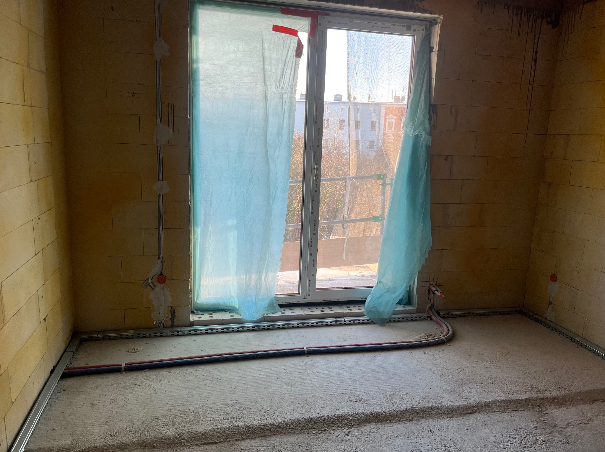 Surowy pokój z instalacją elektryczną w ścianie i rurami w podłodze, okno zasłonięte folią ochronną. Widoczne kable i puszki elektryczne. Stan deweloperski.