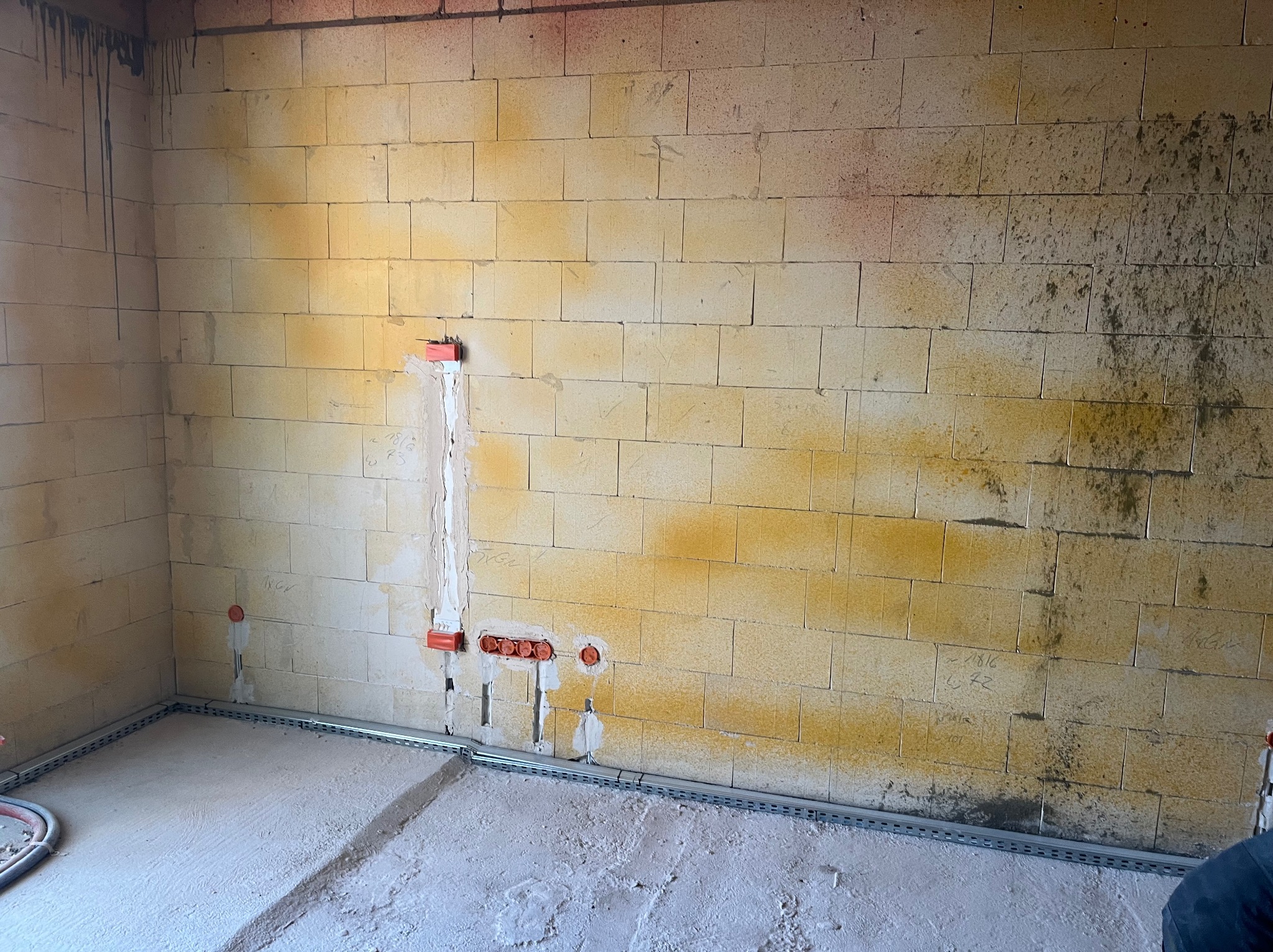 Wnętrze w trakcie prac elektrycznych: widoczne koryta kablowe przy podłodze i puszki instalacyjne w ścianie z betonu komórkowego. Ściany surowe, z widocznymi śladami po budowie.