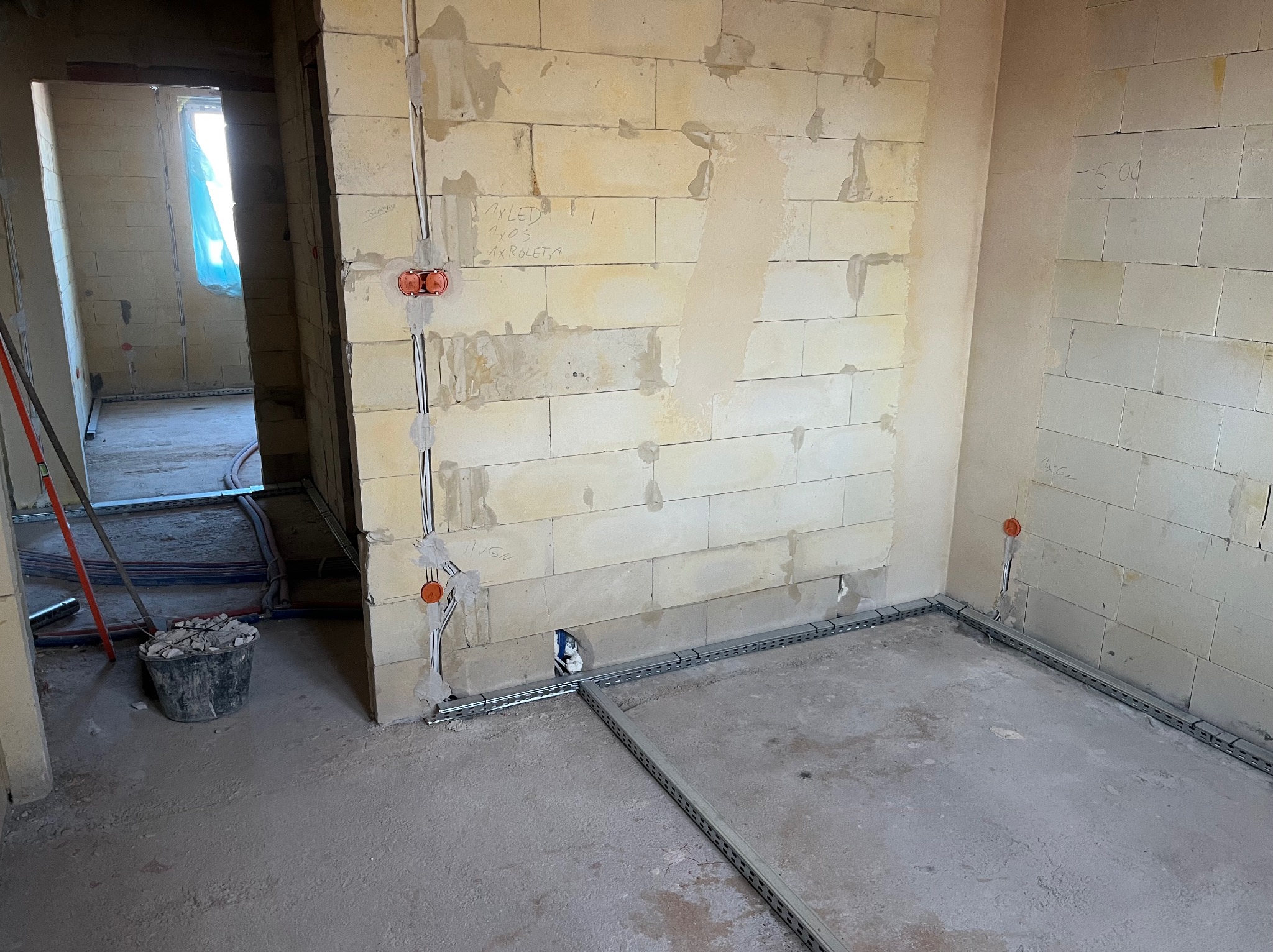 Wnętrze w trakcie instalacji elektrycznej: kable w peszlach, puszki podtynkowe, metalowe korytka kablowe na podłodze. Ściany z bloczków, surowy stan.