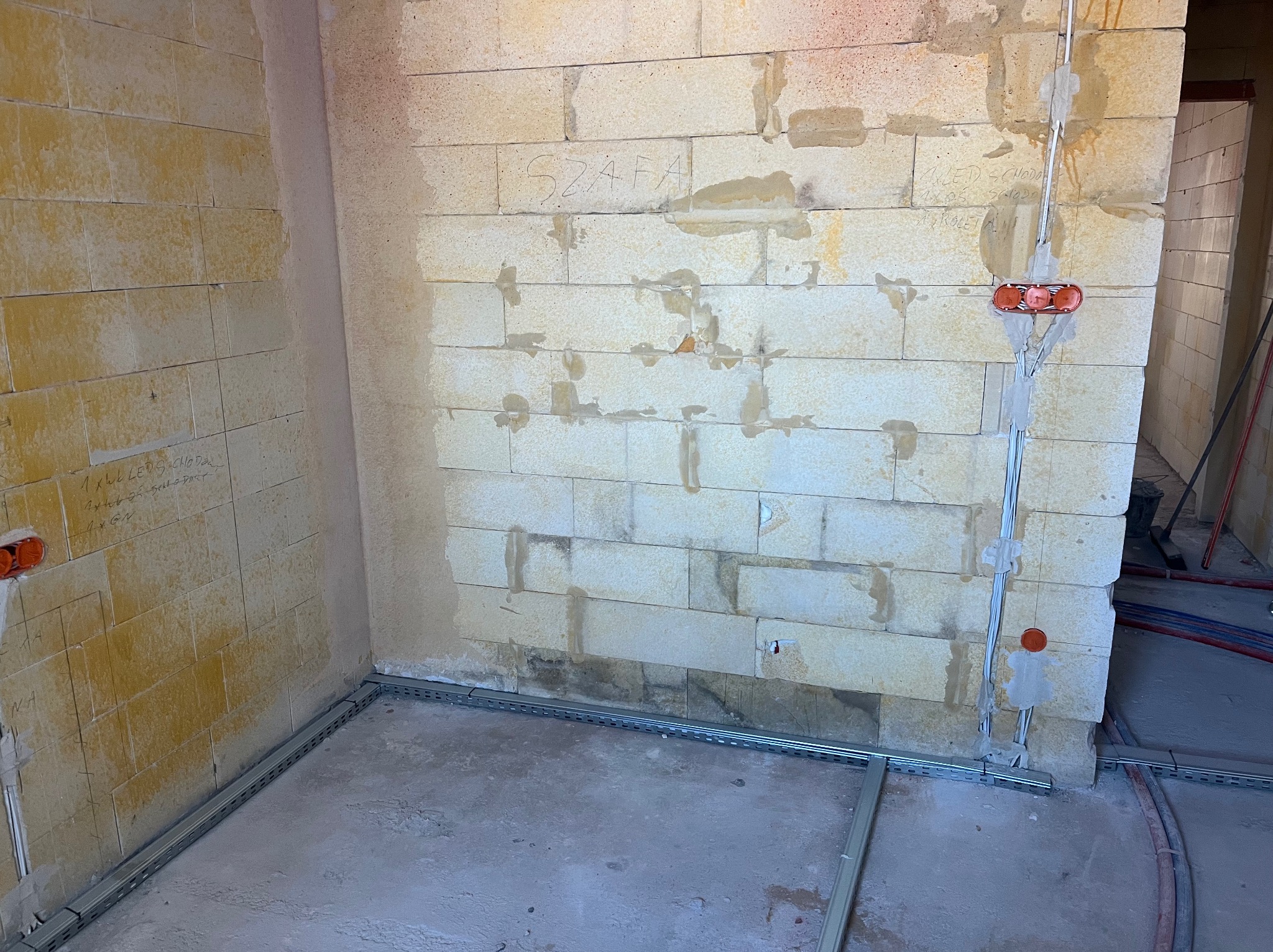 Surowy stan: wnętrze w trakcie prac elektrycznych. Widoczne kable w peszlach, puszki instalacyjne i metalowe korytka kablowe przy ścianach z bloczków.