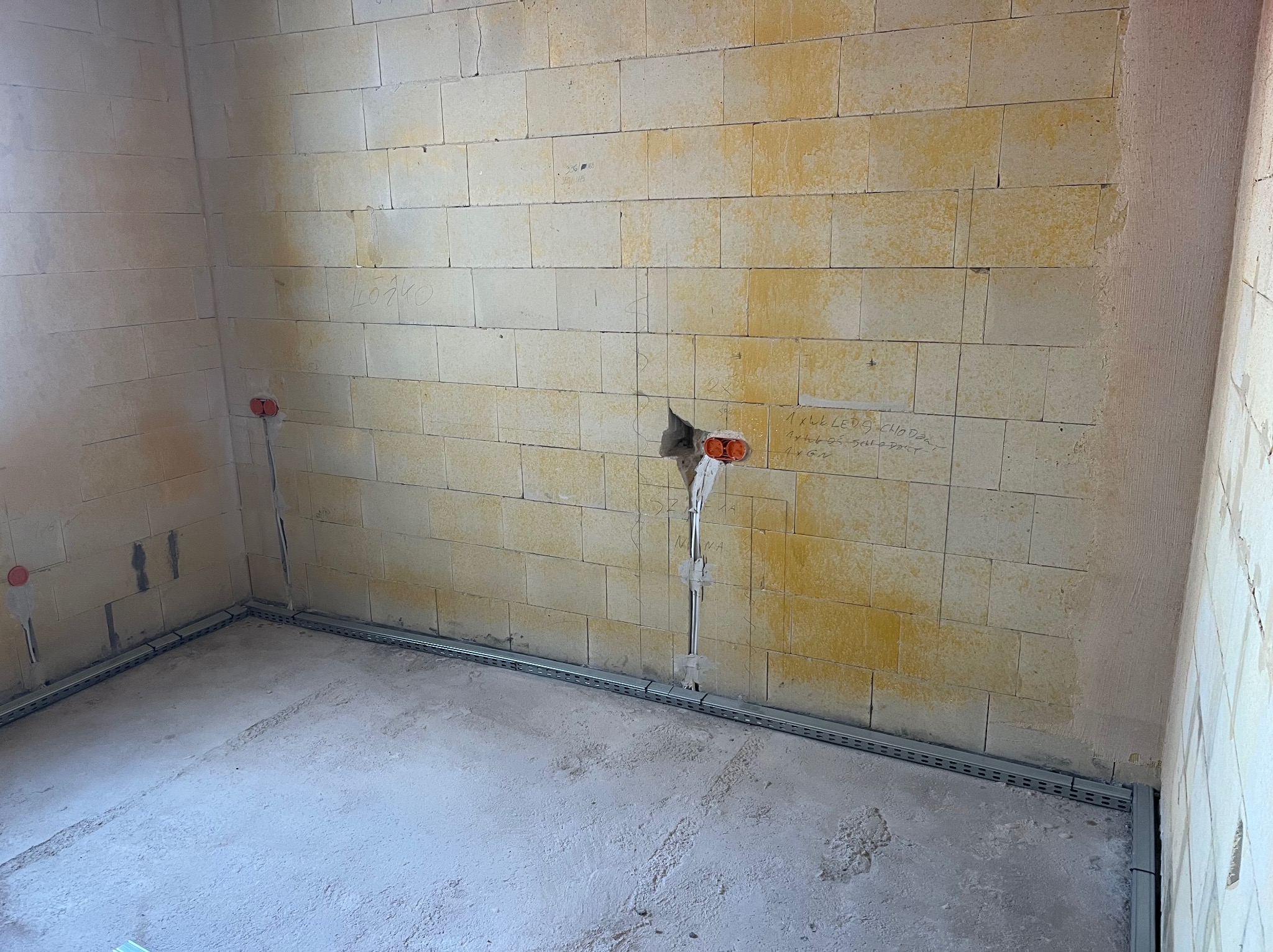 Surowy pokój z instalacją elektryczną: kable w ścianie, puszki podtynkowe i metalowe korytka kablowe wzdłuż ścian. Widoczne napisy na ścianie z bloczków.