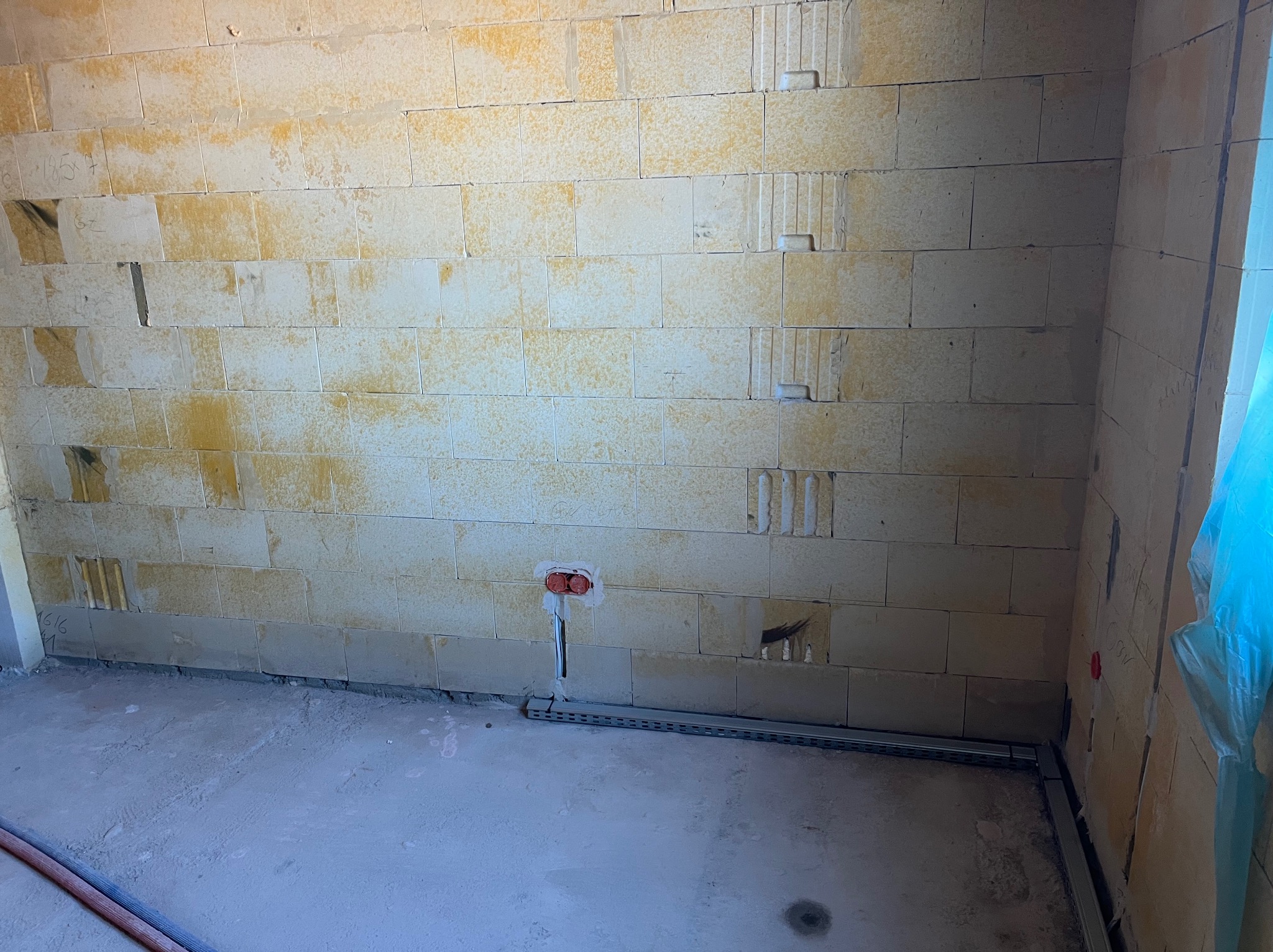 Ściana z pustaków z puszką elektryczną i korytem kablowym przy podłodze, widoczne przewody. Etap prac instalacyjnych w surowym wnętrzu.