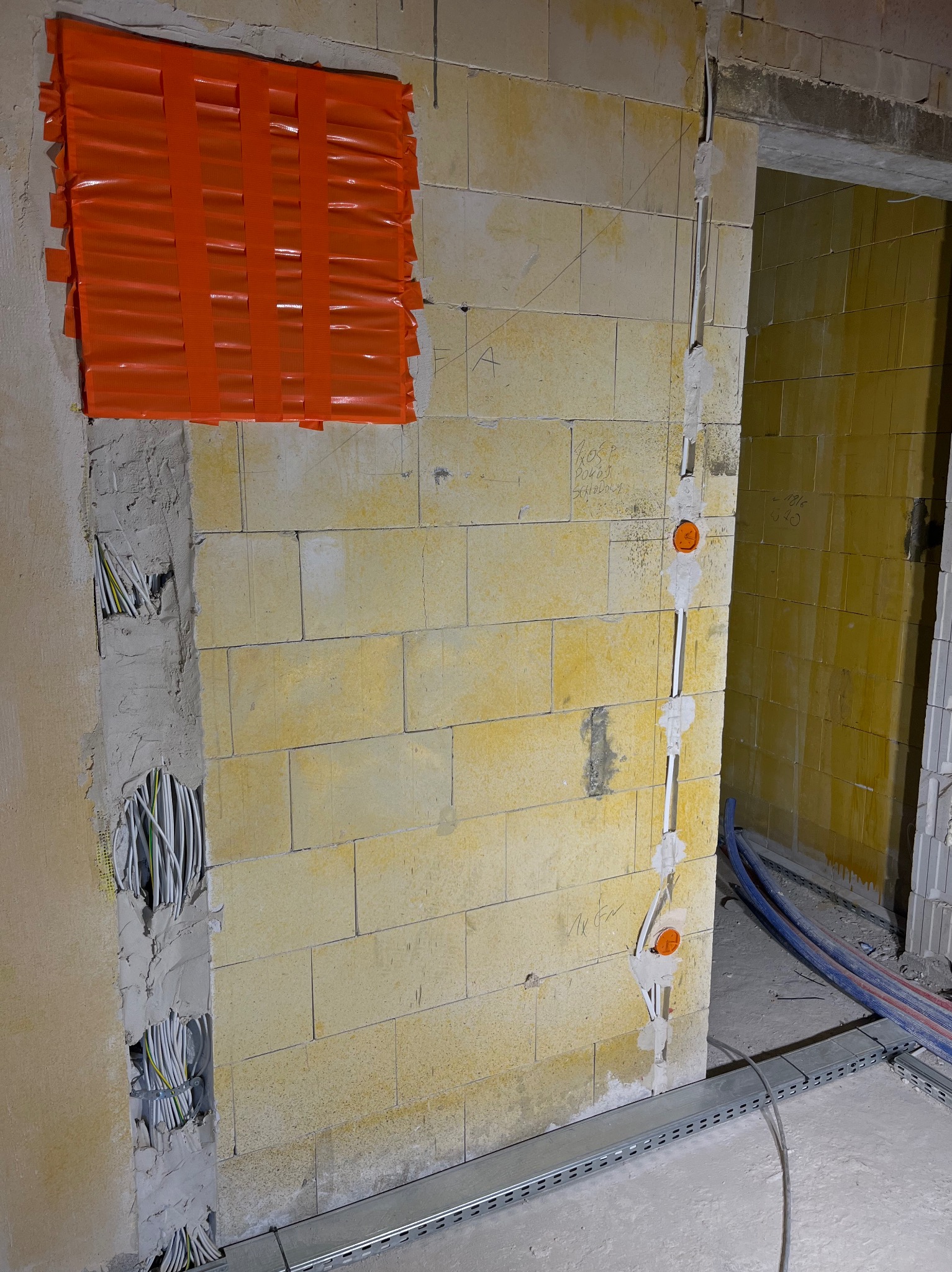 Ściana z instalacją elektryczną w trakcie montażu: kable w bruzdach, zabezpieczone pomarańczową taśmą, puszki elektryczne. Widoczne koryto kablowe przy podłodze.