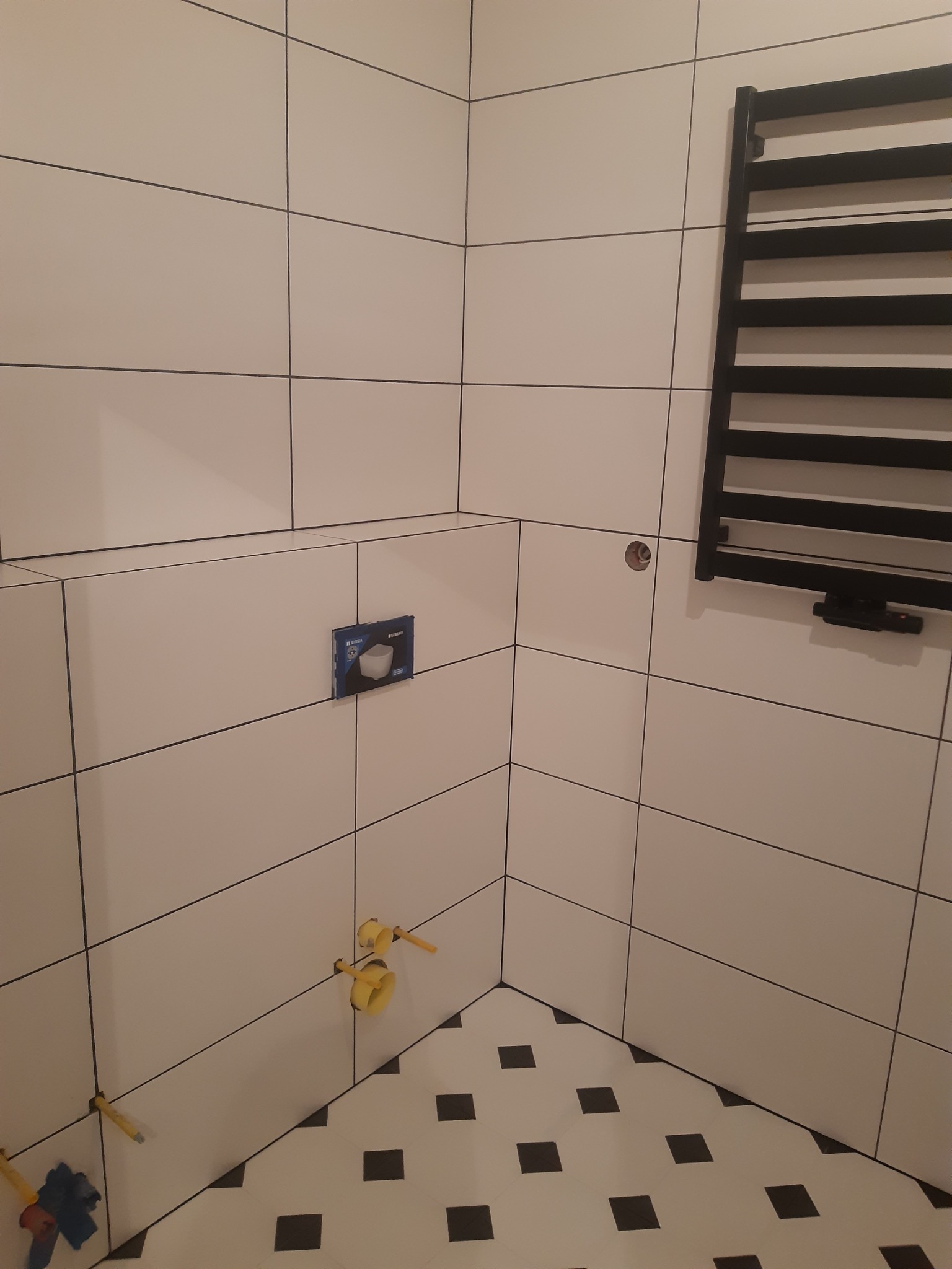 Wykończona kabina prysznicowa z białymi płytkami i czarnymi fugami, czarny grzejnik drabinkowy, podłoga w biało-czarną szachownicę. Widoczne przyłącza wodne i stelaż WC.