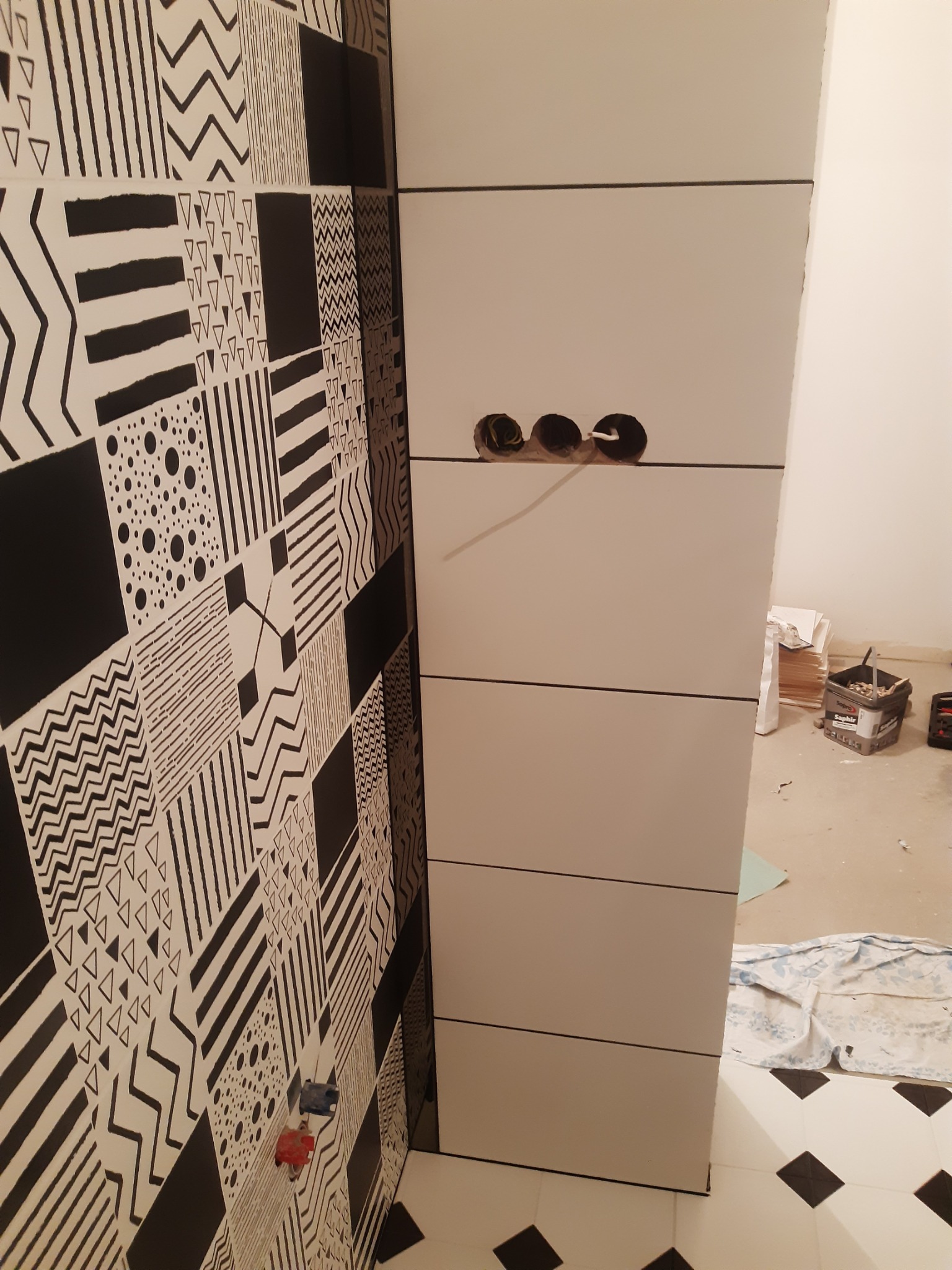 Nowoczesna łazienka w trakcie wykończenia: białe płytki ułożone poziomo, wzorzysta ściana z czarno-białymi motywami, widoczne puszki elektryczne.