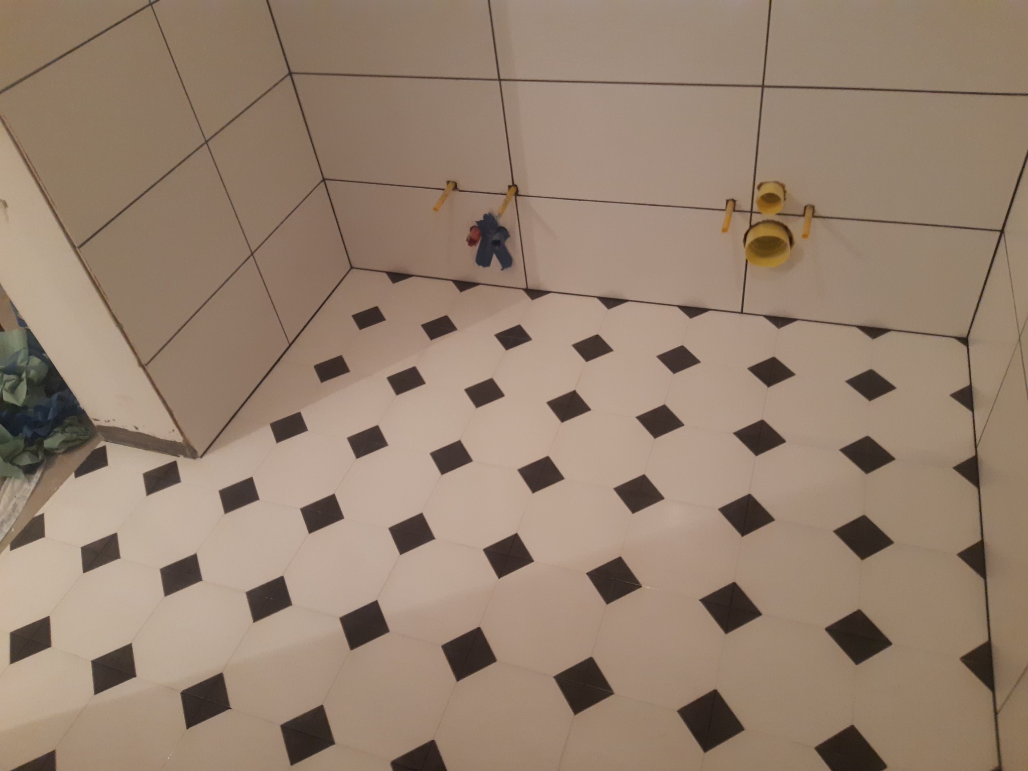 Wykończona podłoga z biało-czarną mozaiką w łazience, białe płytki na ścianach, widoczne elementy instalacji wodnej.