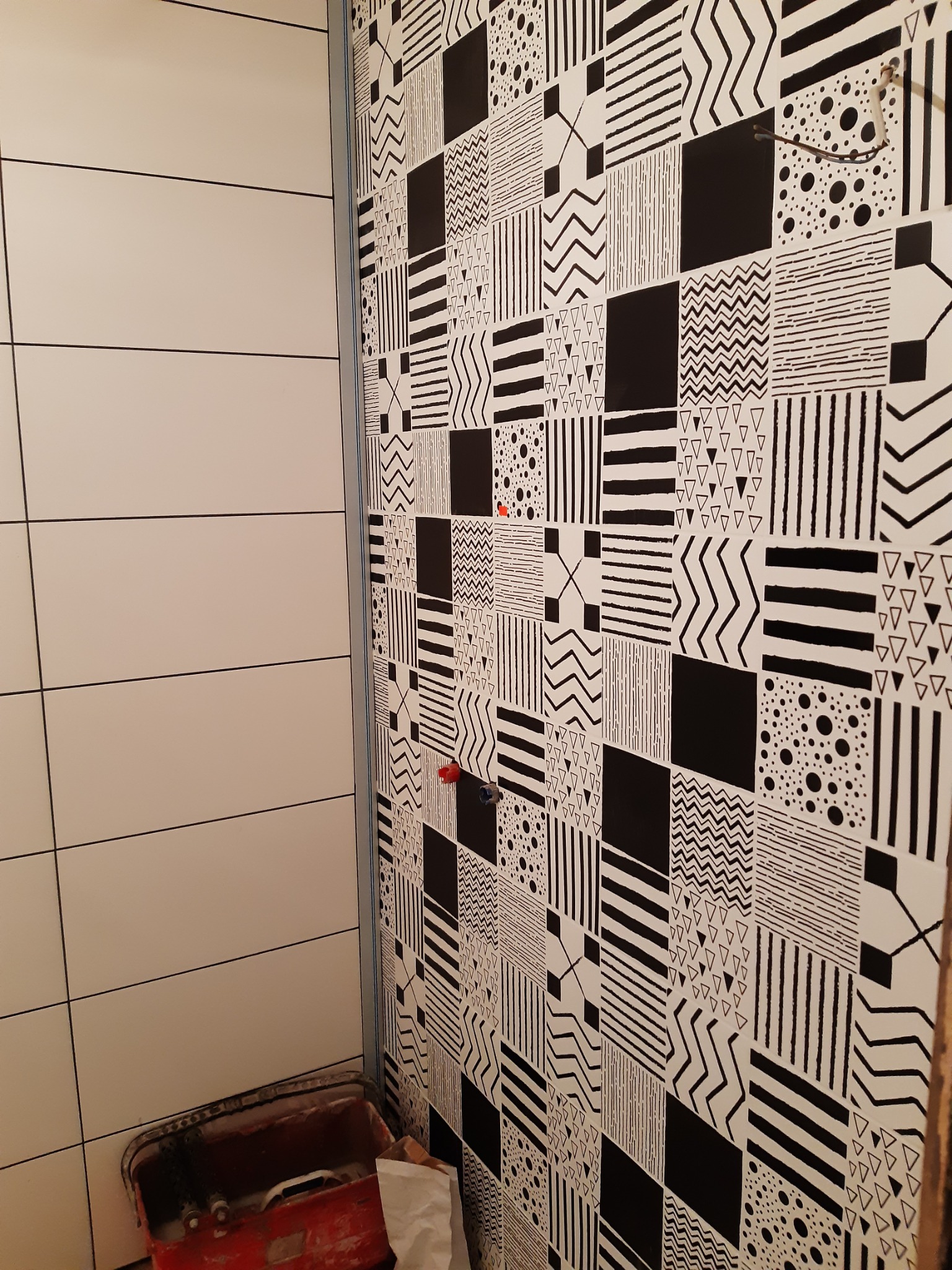 Ściana łazienki wyłożona białymi płytkami i fragment ściany z czarno-białymi płytkami o geometrycznych wzorach. Widoczny fragment czerwonego pojemnika.