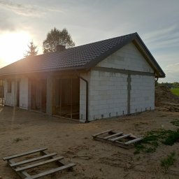 Usługi ogólnobudowlane MasterMaxBud - Budowa domu z białych bloczków w trakcie realizacji. Widoczne zarysy okien i drzwi, szara dachówka, palety drewniane na piaszczystym terenie. Słońce zachodzi za budynkiem.