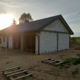 Budowa domu z białych bloczków w trakcie realizacji. Widoczne zarysy okien i drzwi, szara dachówka, palety drewniane na piaszczystym terenie. Słońce zachodzi za budynkiem.