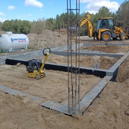 Usługi ogólnobudowlane MasterMaxBud - Fundamenty domu w trakcie budowy: zbrojenie, bloczki betonowe, ubijarka, zbiornik gazu i koparka JCB w tle. Widok na plac budowy w słoneczny dzień.