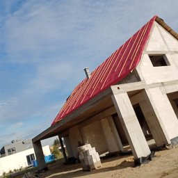Usługi ogólnobudowlane MasterMaxBud - Surowy dom jednorodzinny w trakcie budowy, widok z boku. Czerwona folia i łaty na dachu. Ściany z białych bloczków. W tle błękitne niebo i inne budynki.