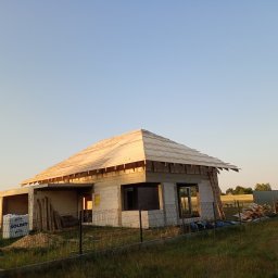 Usługi ogólnobudowlane MasterMaxBud - Budowa domu jednorodzinnego w trakcie realizacji: ściany z bloczków, więźba dachowa, widoczne materiały budowlane, ogrodzenie panelowe i otaczająca zieleń.