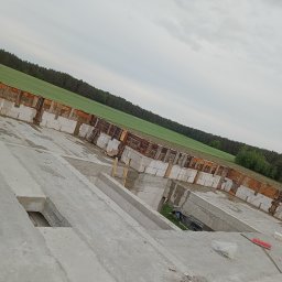 Usługi ogólnobudowlane MasterMaxBud - Surowy fundament budynku z bloczków betonowych i drewnianego szalunku, widok z góry na tle zielonego pola i pochmurnego nieba. Etap budowy.
