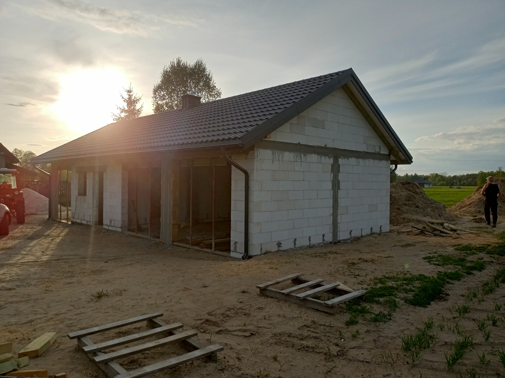Budowa domu z białych bloczków w trakcie realizacji. Widoczne zarysy okien i drzwi, szara dachówka, palety drewniane na piaszczystym terenie. Słońce zachodzi za budynkiem.