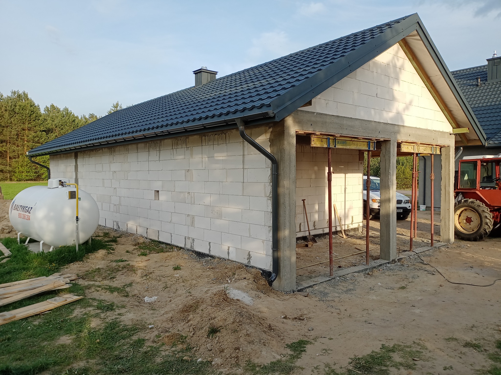 Budowa parterowego domu z białych bloczków, widoczny zbiornik na gaz i czerwony traktor w tle. Etap murowania ścian i przygotowania do wylania stropu.