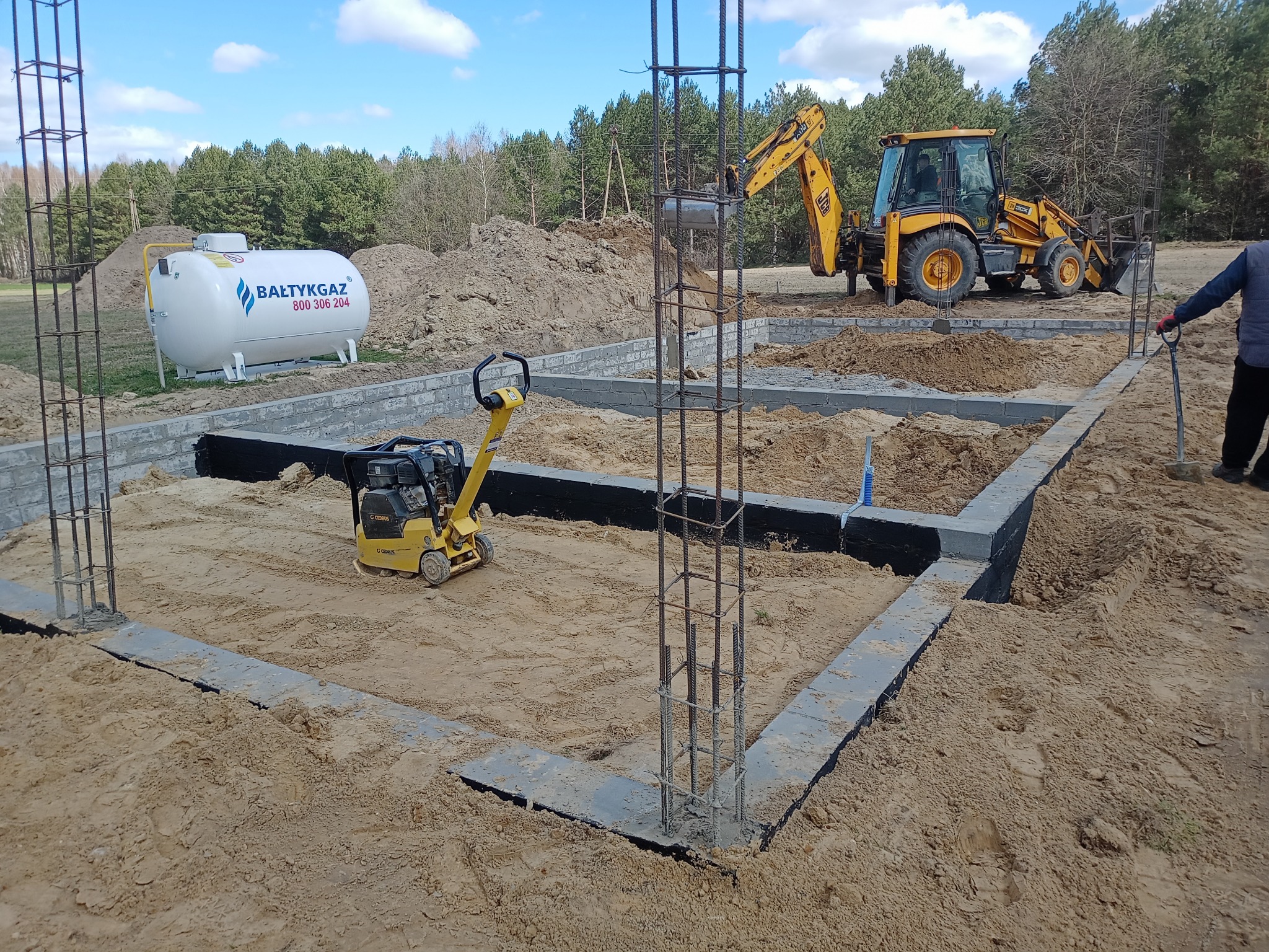 Fundamenty domu w trakcie budowy: zbrojenie, bloczki betonowe, ubijarka, zbiornik gazu i koparka JCB w tle. Widok na plac budowy w słoneczny dzień.