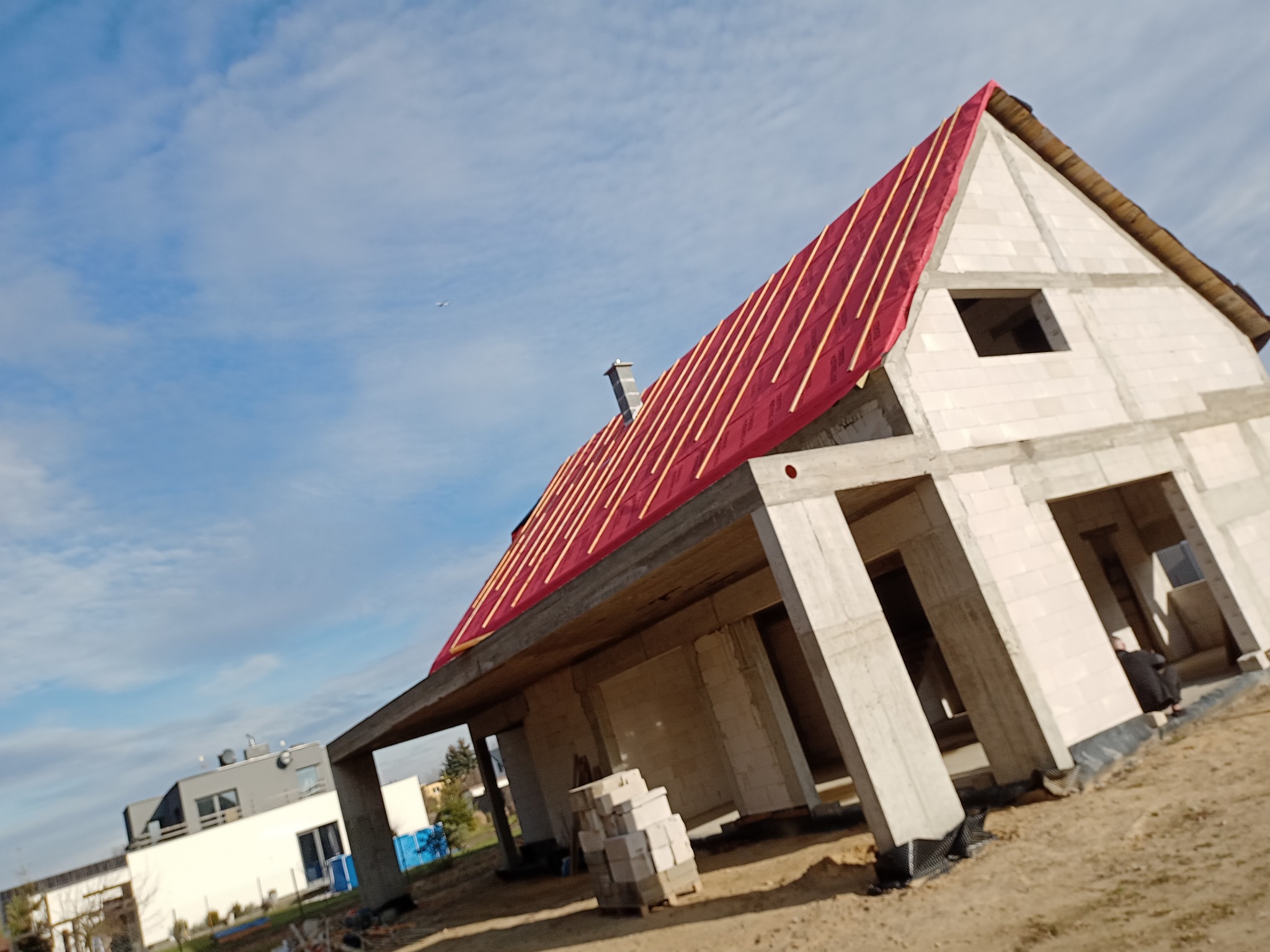 Surowy dom jednorodzinny w trakcie budowy, widok z boku. Czerwona folia i łaty na dachu. Ściany z białych bloczków. W tle błękitne niebo i inne budynki.