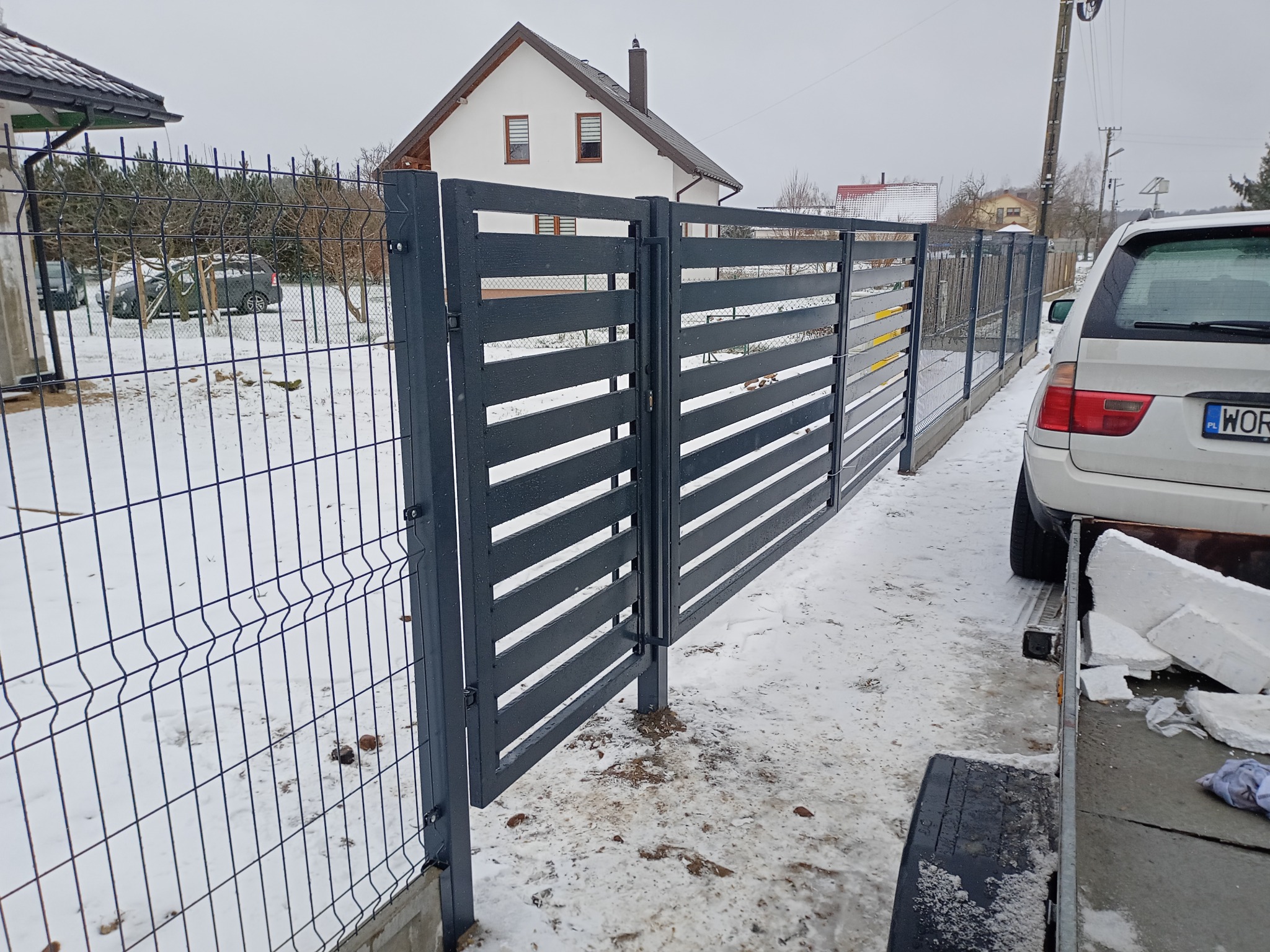 Nowoczesne, grafitowe ogrodzenie panelowe z poziomymi przęsłami i furtką, połączone z siatką. W tle dom jednorodzinny i śnieżny krajobraz.