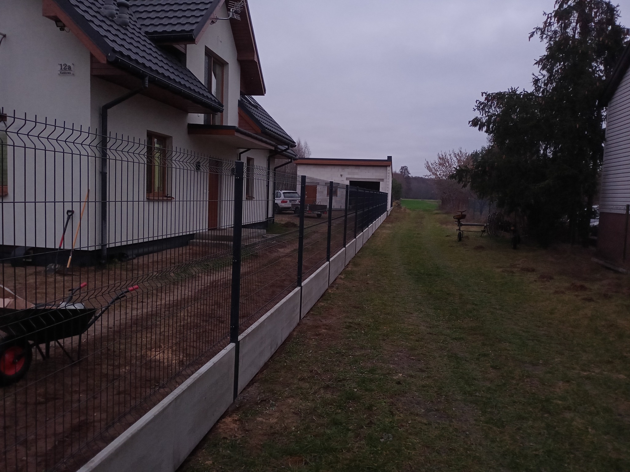 Nowoczesne ogrodzenie panelowe z podmurówką betonową, widok z boku. Na pierwszym planie taczka i narzędzia ogrodnicze. W tle dom z ciemnym dachem i budynek gospodarczy.