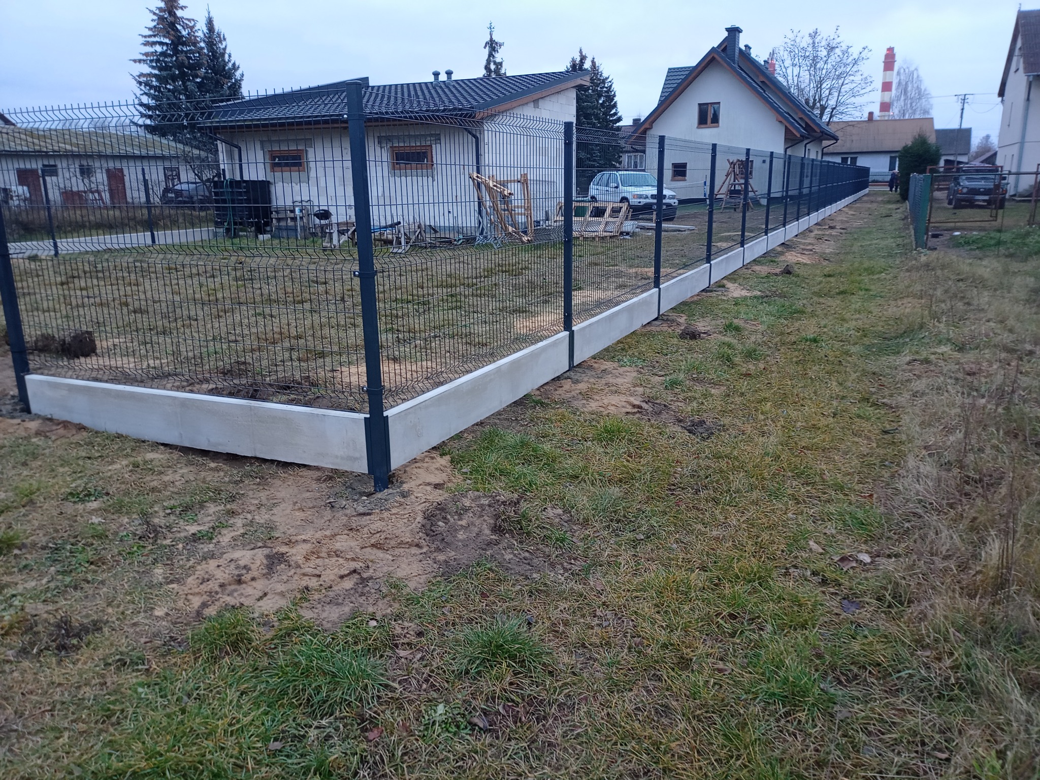 Nowoczesne ogrodzenie panelowe z podmurówką betonową, widok z boku na tle zabudowań. Słupki w kolorze grafitowym. Widoczne elementy montażowe.