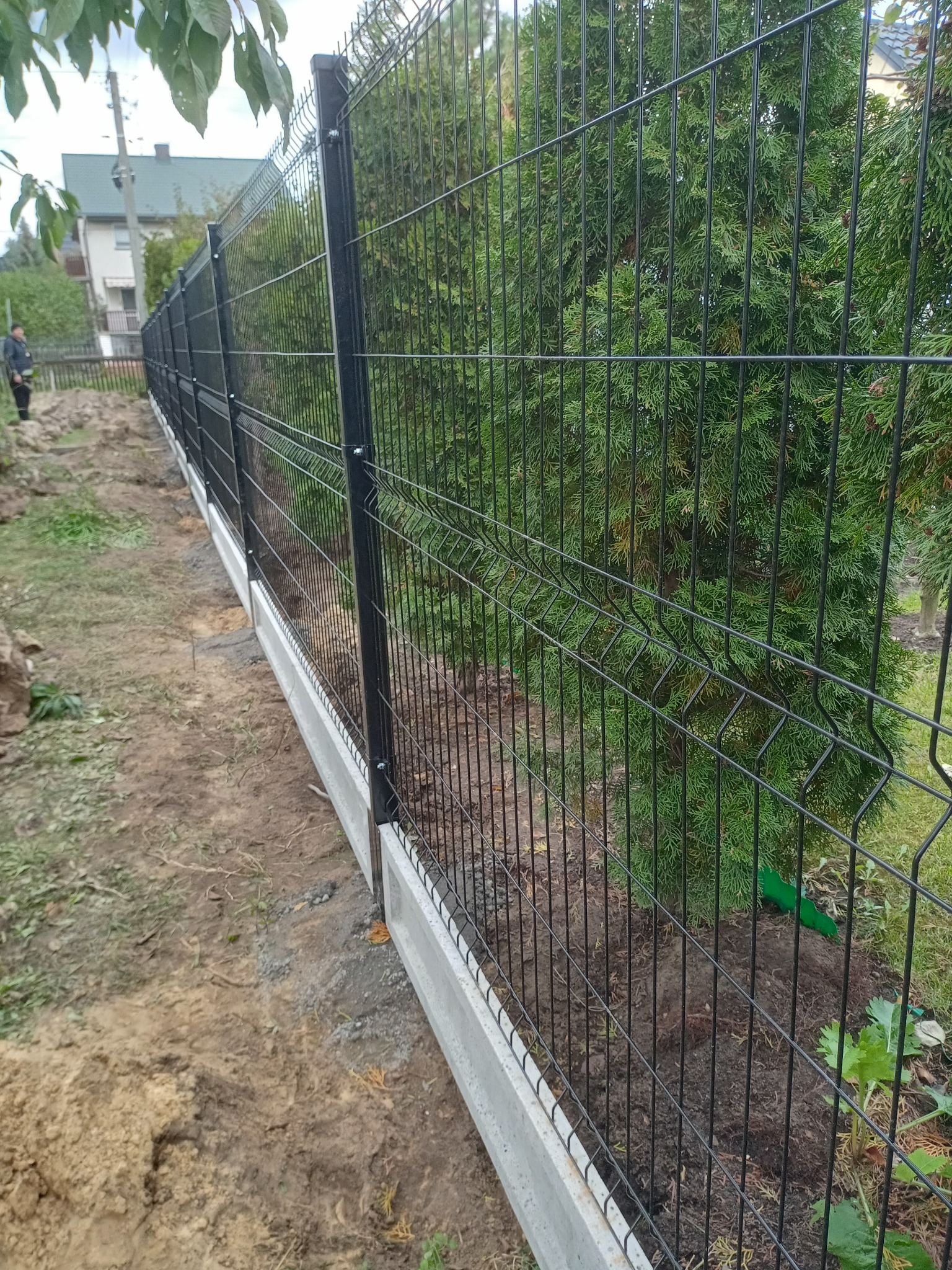 Nowoczesne ogrodzenie panelowe z podmurówką betonową, widok z boku na tle zieleni. Świeżo usypana ziemia wzdłuż ogrodzenia. Czarny metal kontrastuje z szarą podmurówką.