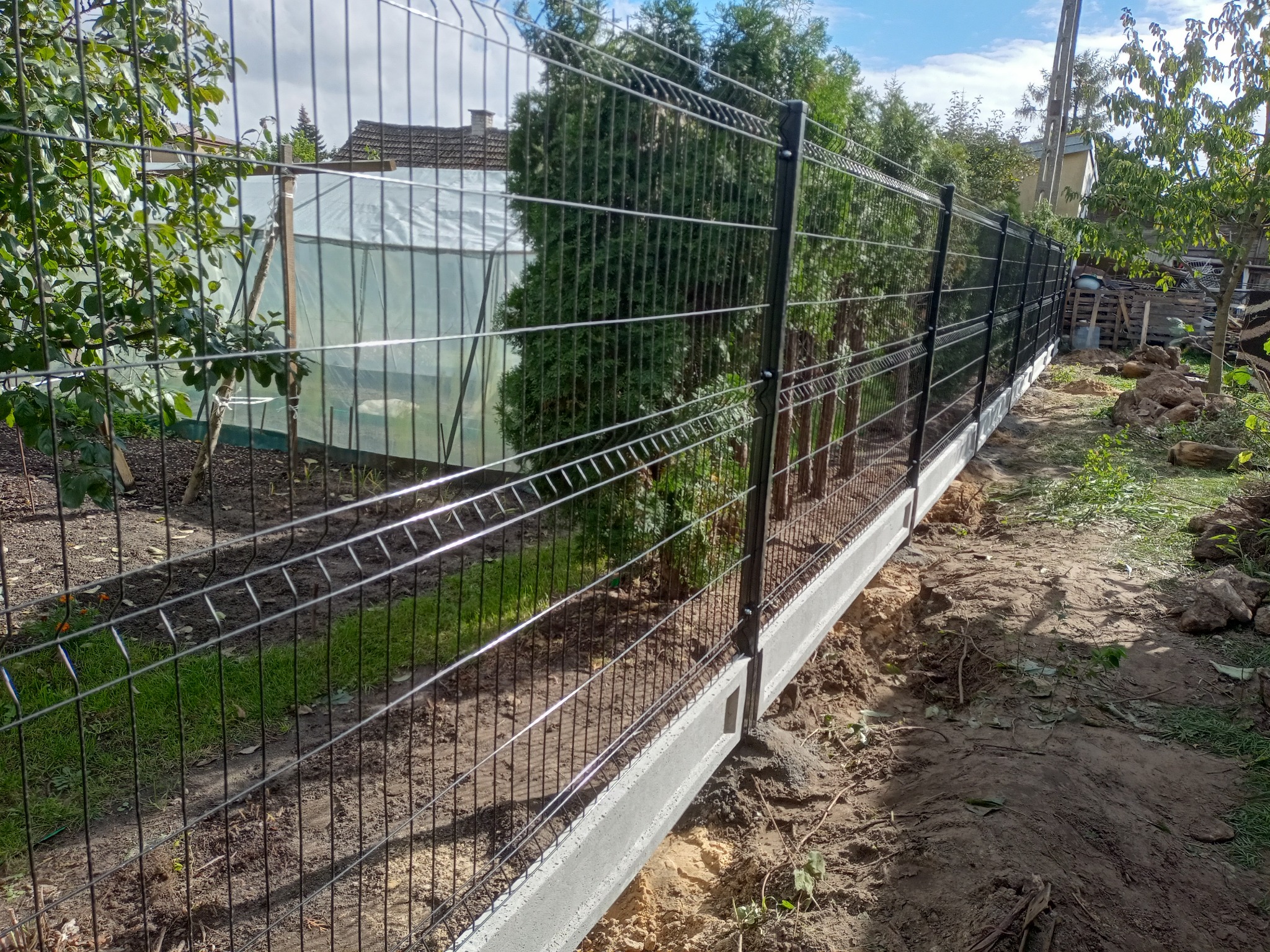 Nowoczesne ogrodzenie panelowe z podmurówką betonową na tle zieleni ogrodowej i błękitnego nieba. Widoczne metalowe panele i słupki.