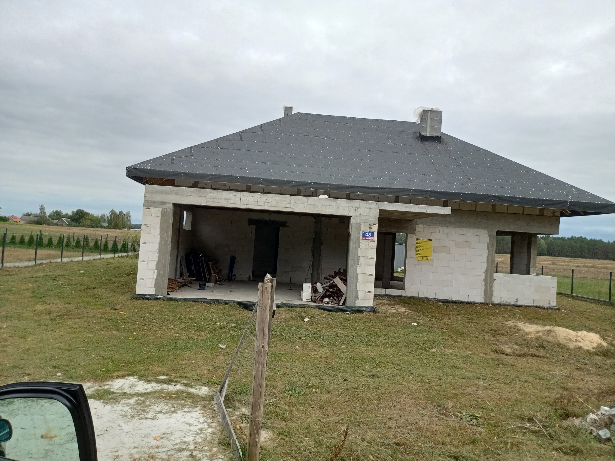 Surowy dom jednorodzinny w trakcie budowy, z widoczną konstrukcją dachu pokrytą papą, niezadaszony garaż, ściany z bloczków i numer domu na ścianie.