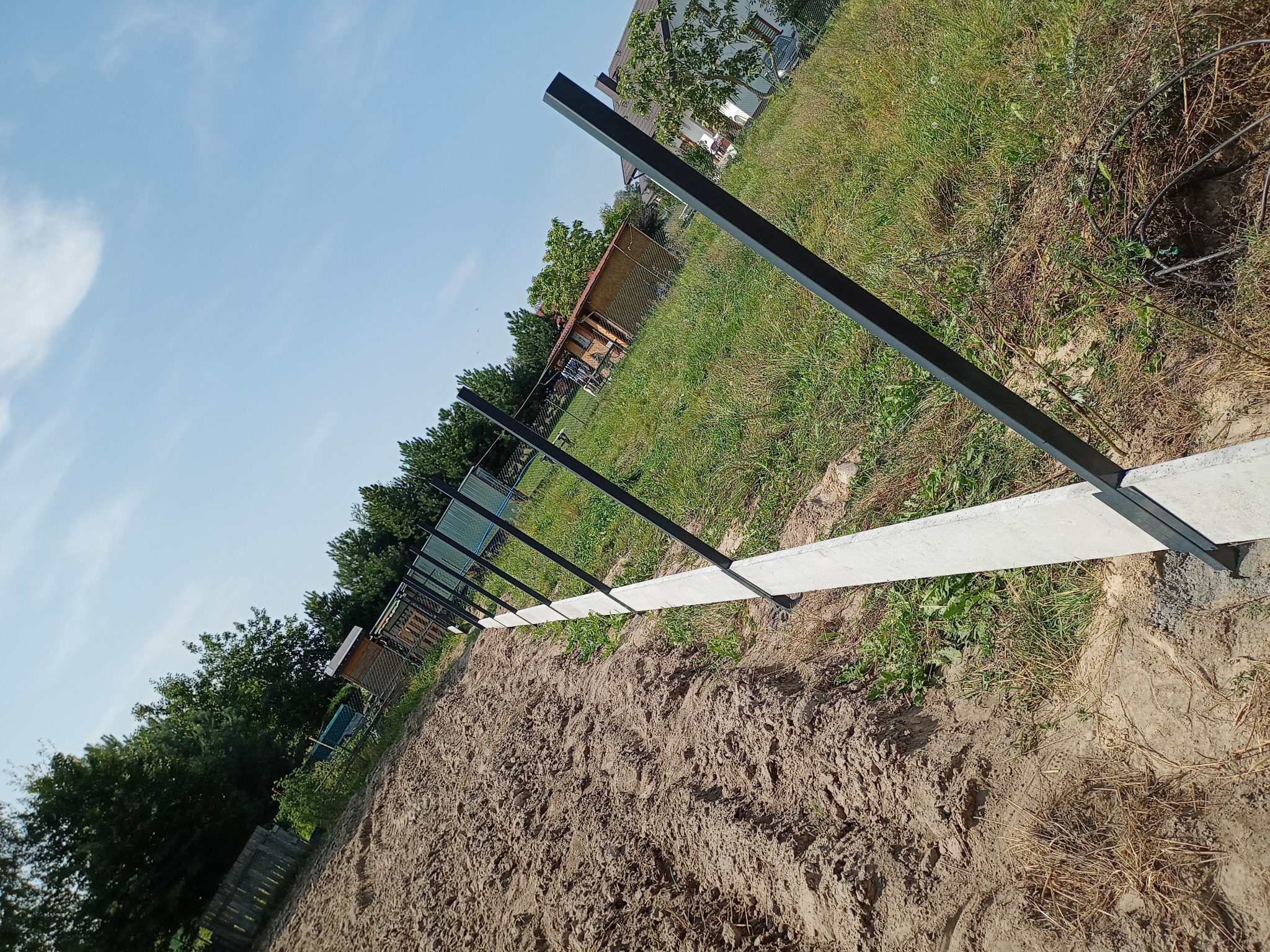 Fundament pod ogrodzenie z metalowymi słupkami w ziemi. Widok na betonową podmurówkę i rząd słupków na tle zieleni i błękitnego nieba.
