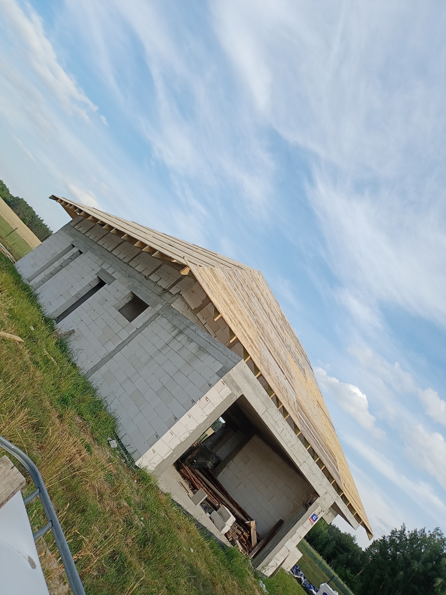 Budowa domu z betonu komórkowego w trakcie realizacji, widok z dołu na ściany i konstrukcję dachu na tle błękitnego nieba z chmurami.