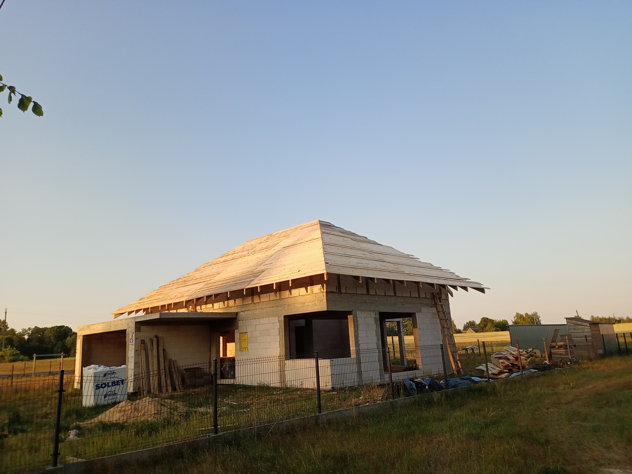 Budowa domu jednorodzinnego w trakcie realizacji: ściany z bloczków, więźba dachowa, widoczne materiały budowlane, ogrodzenie panelowe i otaczająca zieleń.