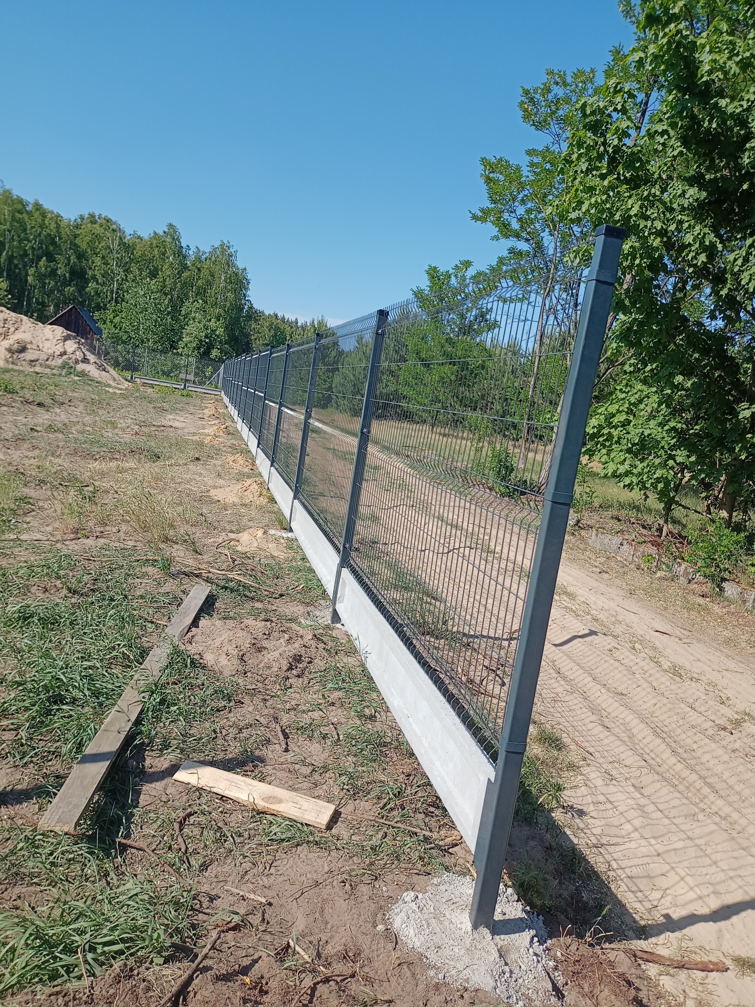 Długi, szary panel ogrodzeniowy na betonowej podmurówce, z metalowymi słupkami, ciągnący się wzdłuż polnej drogi, z drzewami i błękitnym niebem w tle.