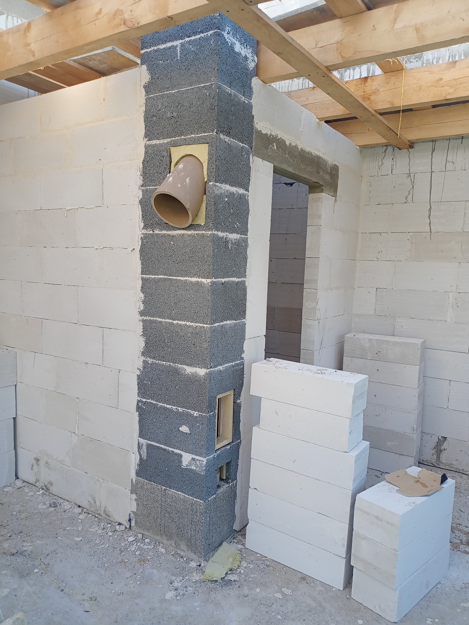 Wnętrze w trakcie budowy: białe bloczki, fragment szarego komina z rurą wentylacyjną, drewniana konstrukcja stropu. Widoczne materiały budowlane i surowy stan wykończenia.