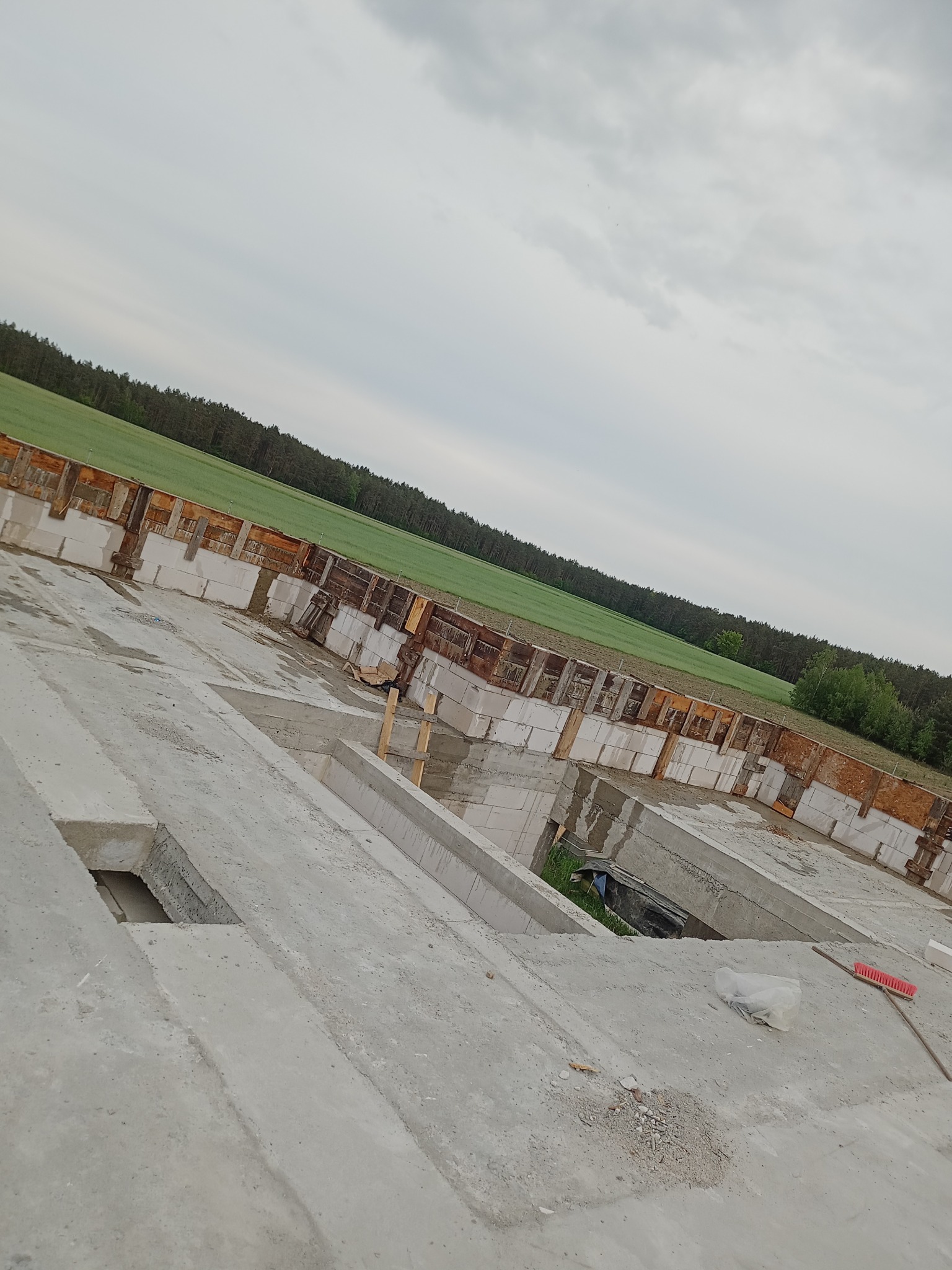 Surowy fundament budynku z bloczków betonowych i drewnianego szalunku, widok z góry na tle zielonego pola i pochmurnego nieba. Etap budowy.