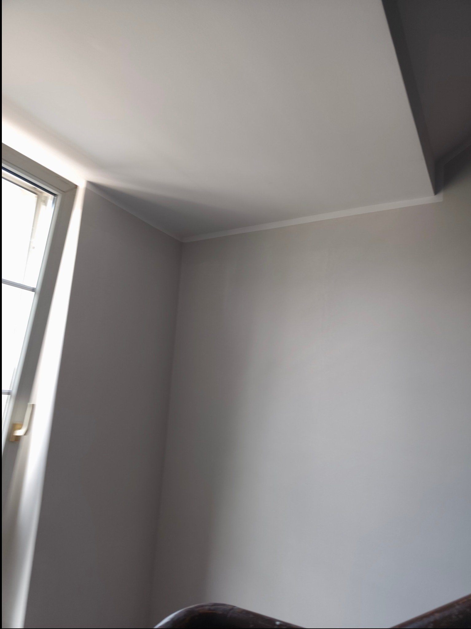 Jasne, świeżo pomalowane wnętrze z oknem po lewej stronie, ukazujące gładką powierzchnię ściany i sufitu w odcieniach bieli i szarości. Delikatne światło dzienne.