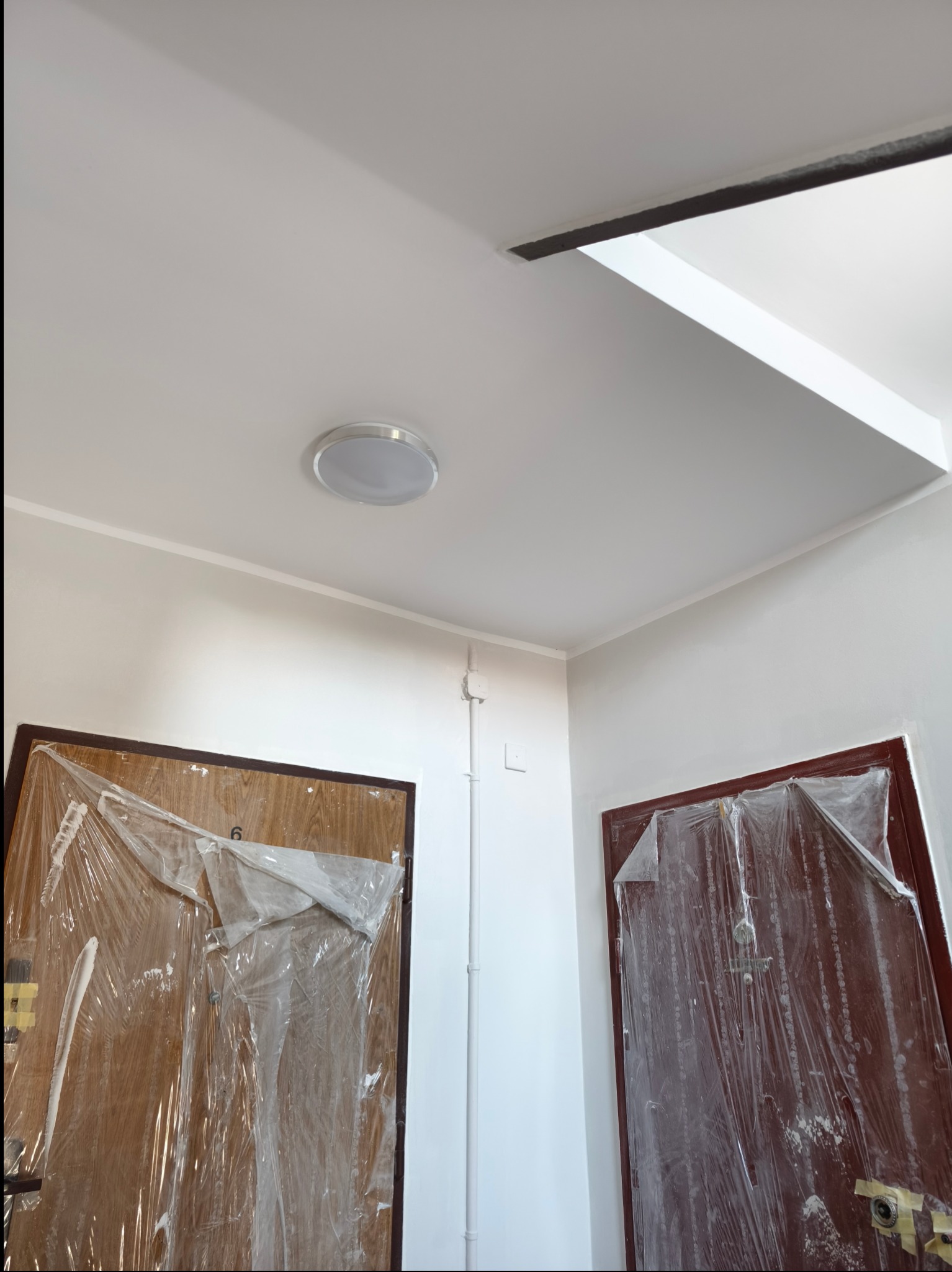 Białe ściany i sufit po malowaniu, zabezpieczone folią drzwi, widoczna lampa sufitowa i listwa dekoracyjna. Świeżo odmalowane wnętrze.