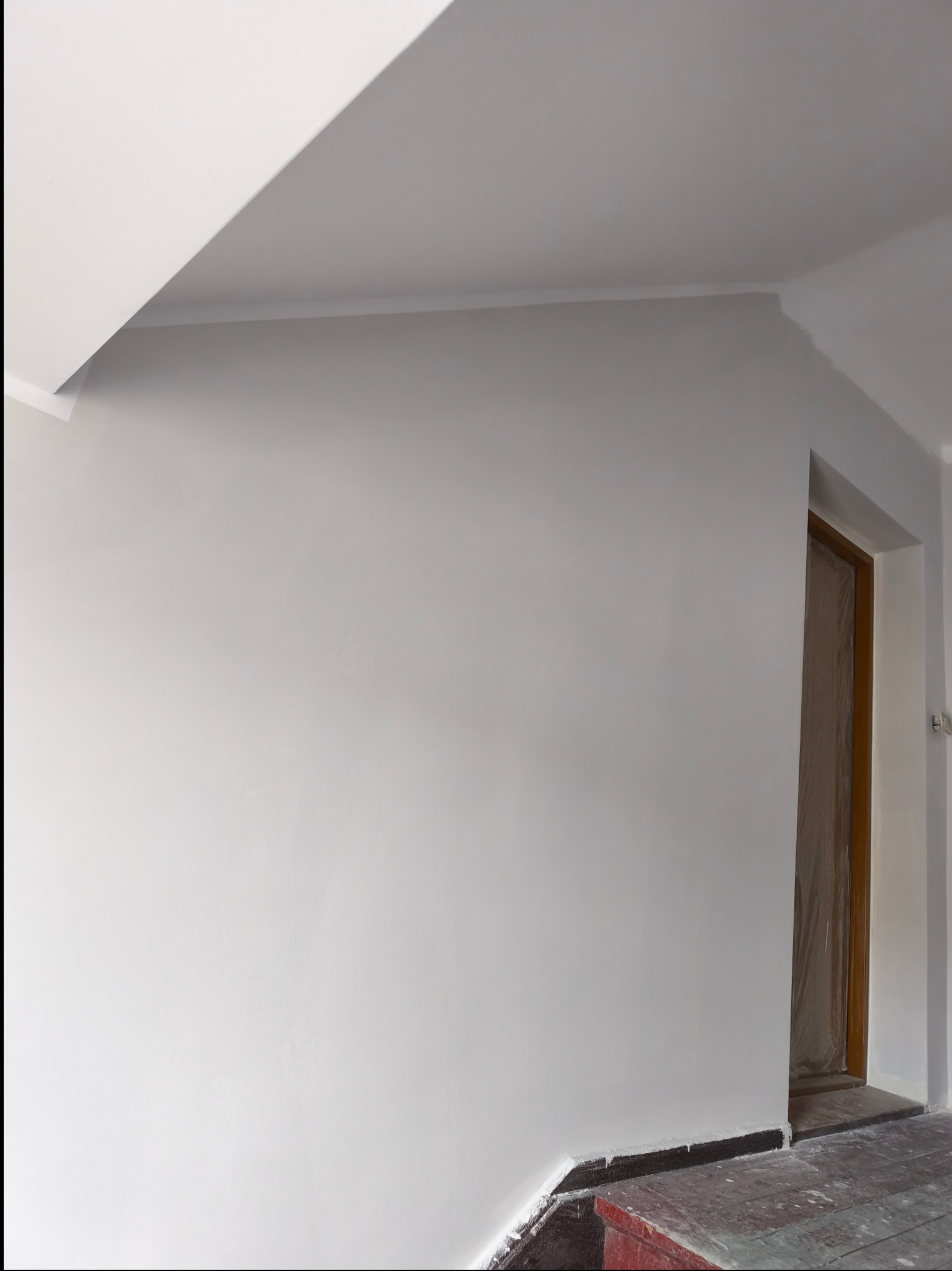 Świeżo pomalowana na szaro ściana z listwą przypodłogową i sufitem. Drzwi zabezpieczone folią malarską. Widoczne ślady farby na podłodze.