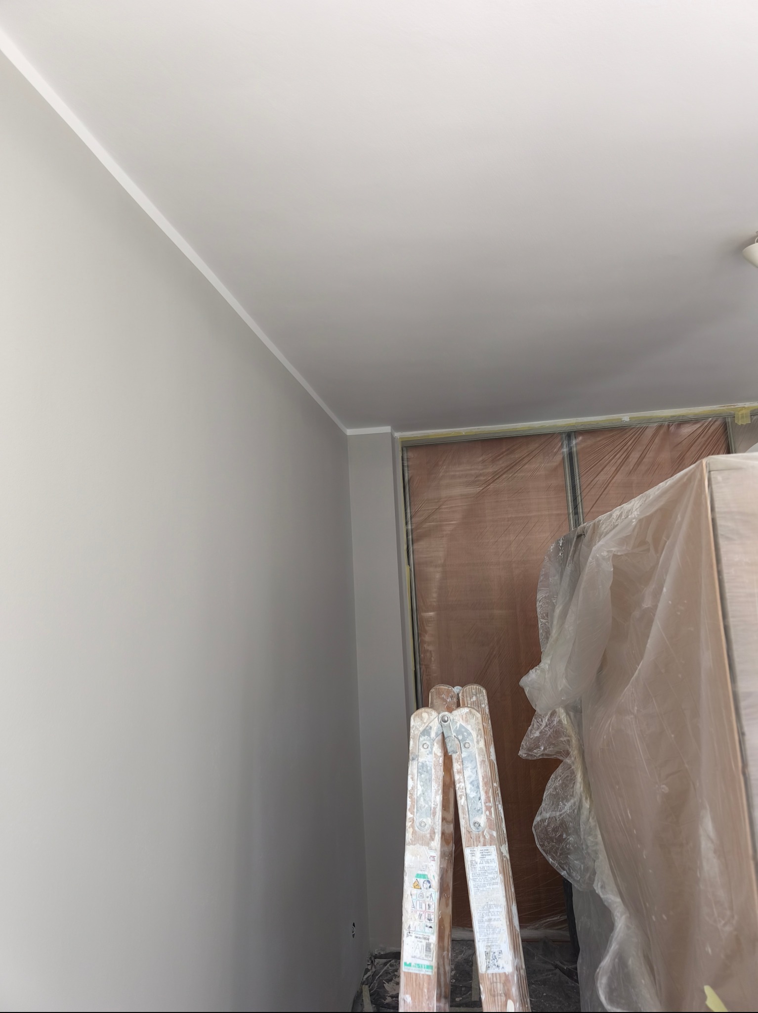 Świeżo pomalowane na szaro wnętrze z białą listwą sufitową. Drabina stoi obok zabezpieczonej folią szafy wnękowej. Widoczny fragment sufitu i ściany.