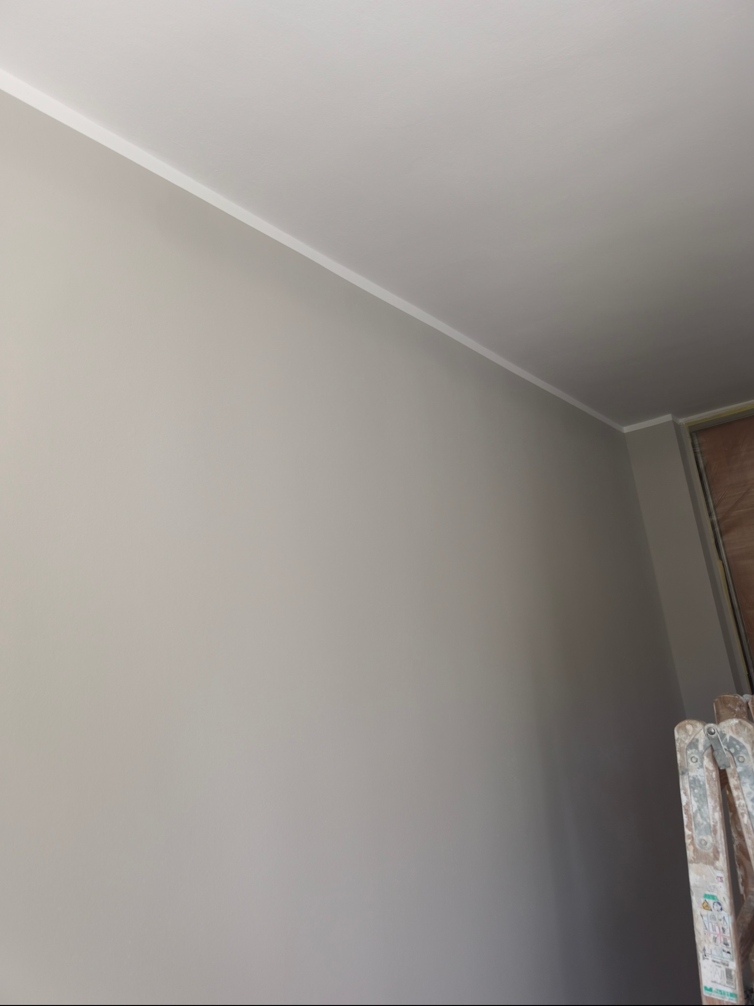 Ściana po malowaniu na szaro, z widoczną białą listwą przysufitową i fragmentem drabiny malarskiej w prawym dolnym rogu. Gładka powierzchnia.