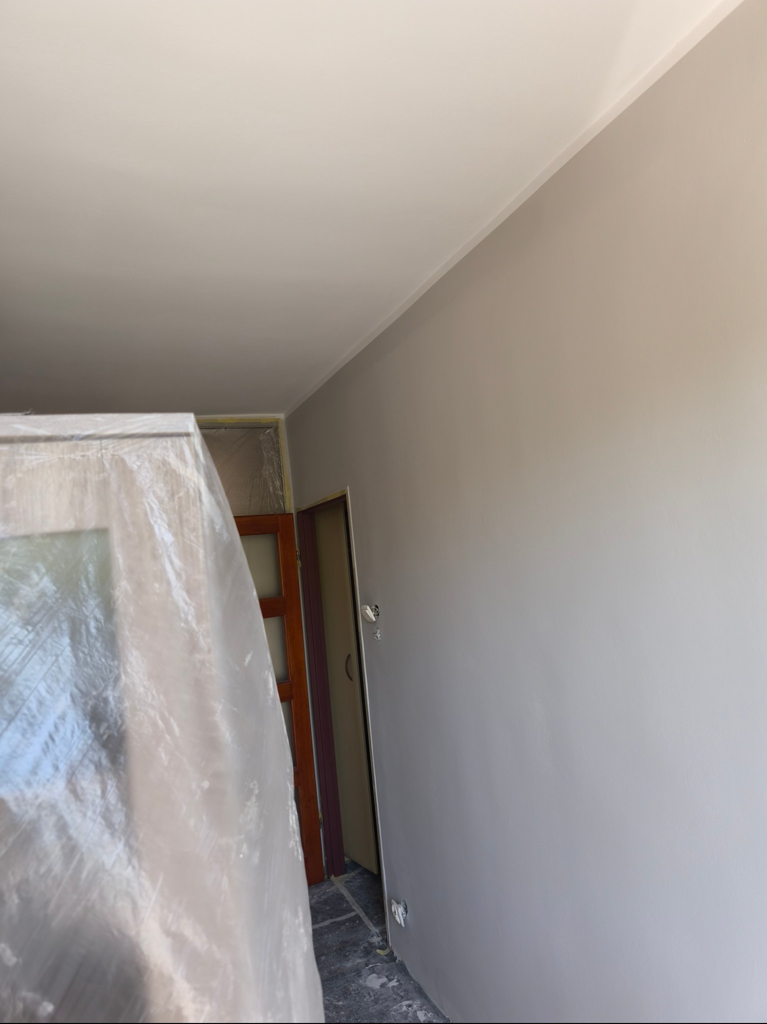 Ściana po malowaniu na szaro, z zabezpieczonymi drzwiami folią malarską. Widoczny fragment sufitu i listwa. Ujęcie z dołu, minimalny kadr.