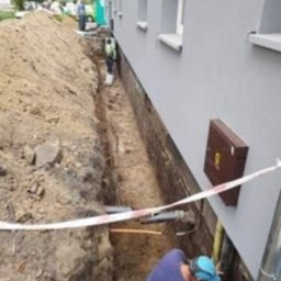 Radosław Głowacz - Wykop wzdłuż ściany budynku z widocznymi rurami i pracownikiem w wykopie, zabezpieczony taśmą ostrzegawczą. Widoczny fragment elewacji budynku.