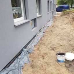 Radosław Głowacz - Jasnoszara elewacja budynku po malowaniu, zabezpieczona folią. Na ziemi wiadro z farbą i pokrywka. W tle widoczny fragment niebieskiego samochodu.