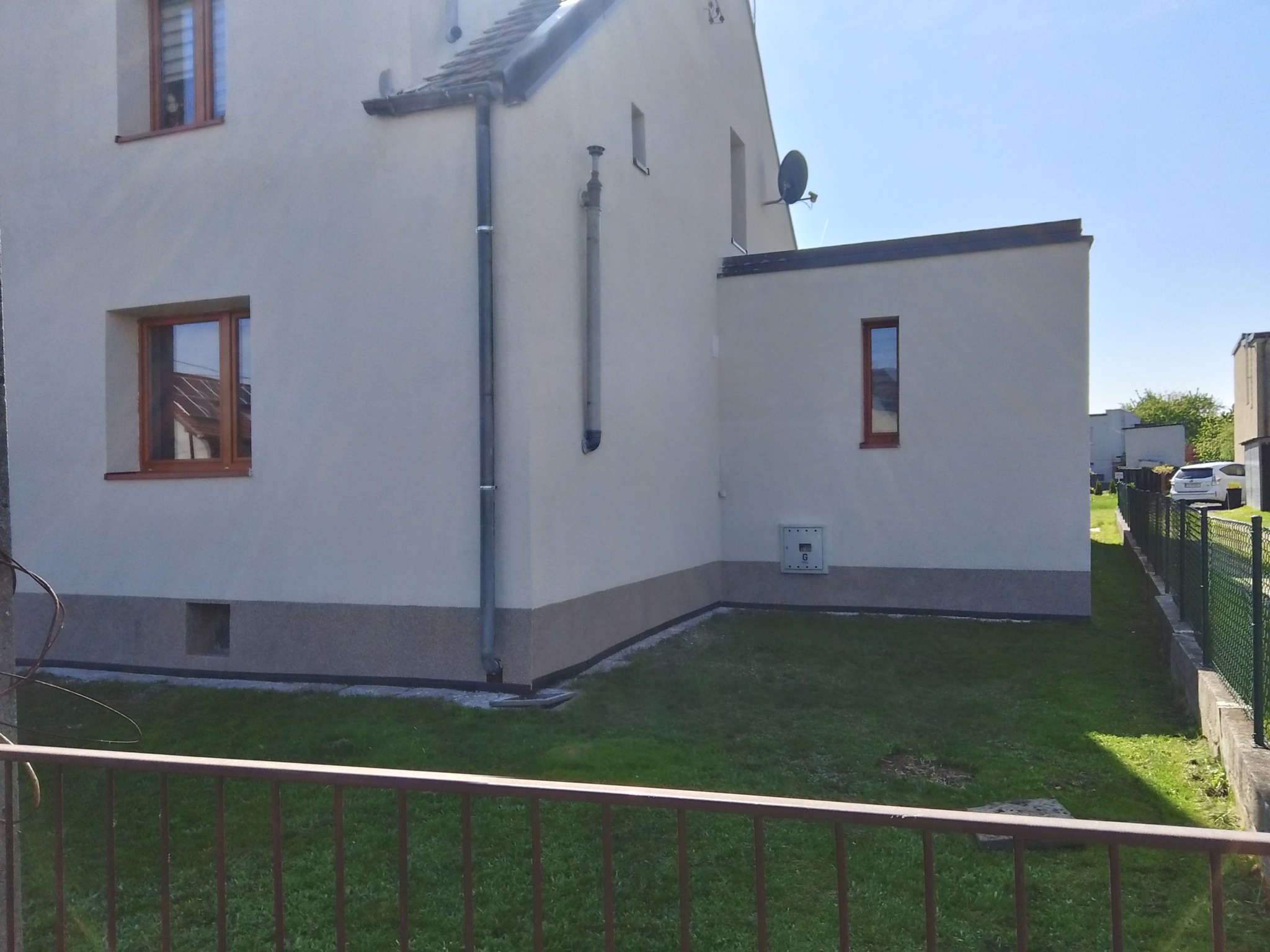 Odnowiona elewacja domu jednorodzinnego z brązowymi ramami okiennymi, widok zza metalowego płotu na trawnik i budynek z anteną satelitarną.