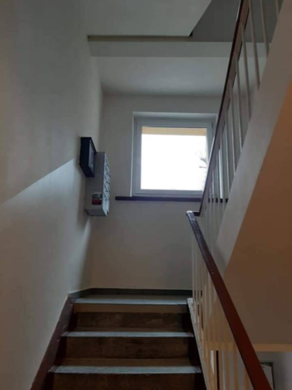 Klatka schodowa w bloku z szarą ścianą, brązowymi schodami i białymi barierkami. Na ścianie skrzynki pocztowe. Okno w tle. Widok z dołu.