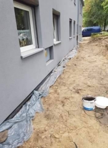 Jasnoszara elewacja budynku po malowaniu, zabezpieczona folią. Na ziemi wiadro z farbą i pokrywka. W tle widoczny fragment niebieskiego samochodu.