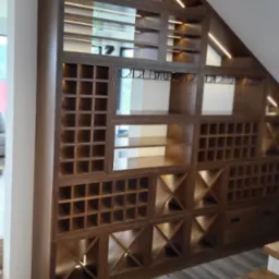 Drewniana zabudowa pod schodami z podświetleniem LED, tworząca regał z przegródkami na wino i szklanki. Widoczne schody z oświetleniem.