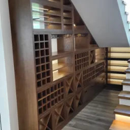 Drewniana szafa wnękowa pod schodami z oświetleniem LED, na wino i inne przedmioty. Widoczna podłoga z drewnopodobnych płytek i fragment schodów.